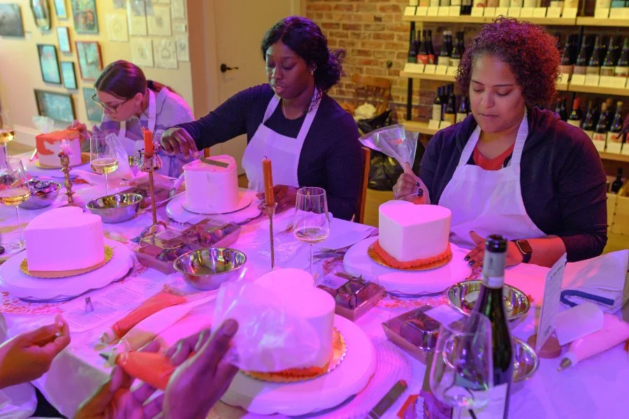 Three women decorating cakes at a table in a cozy, well-lit room with wine bottles on shelves in the background.