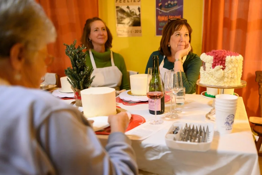 Three women seated at a decorated dining table with a birthday cake, wine, glasses, and holiday decorations, celebrating a special occasion in a cozy, warmly lit room.