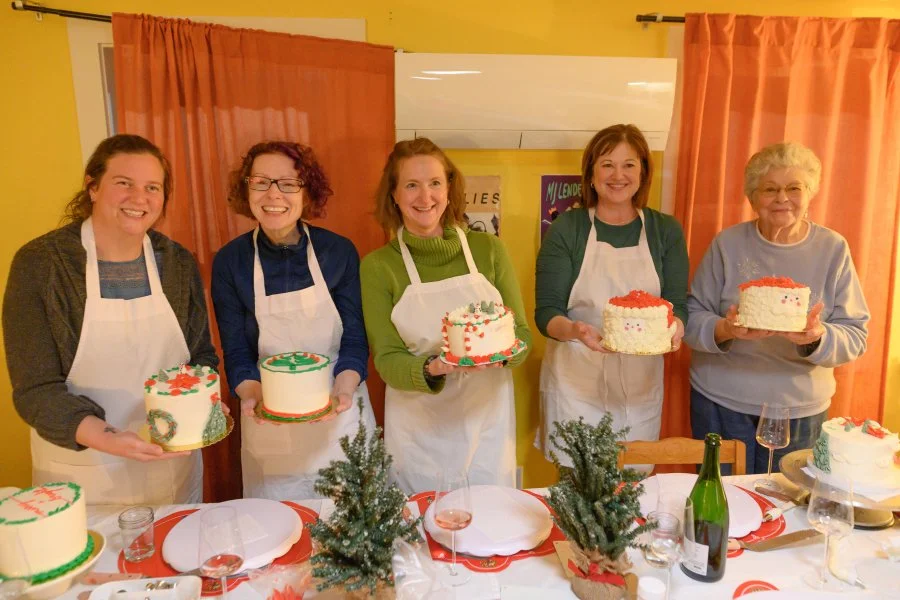 Five women standing behind a table with birthday cakes, holding cakes and smiling. The table is decorated with small Christmas trees, plates, glasses, and a bottle of wine. The room has yellow walls and pink curtains.