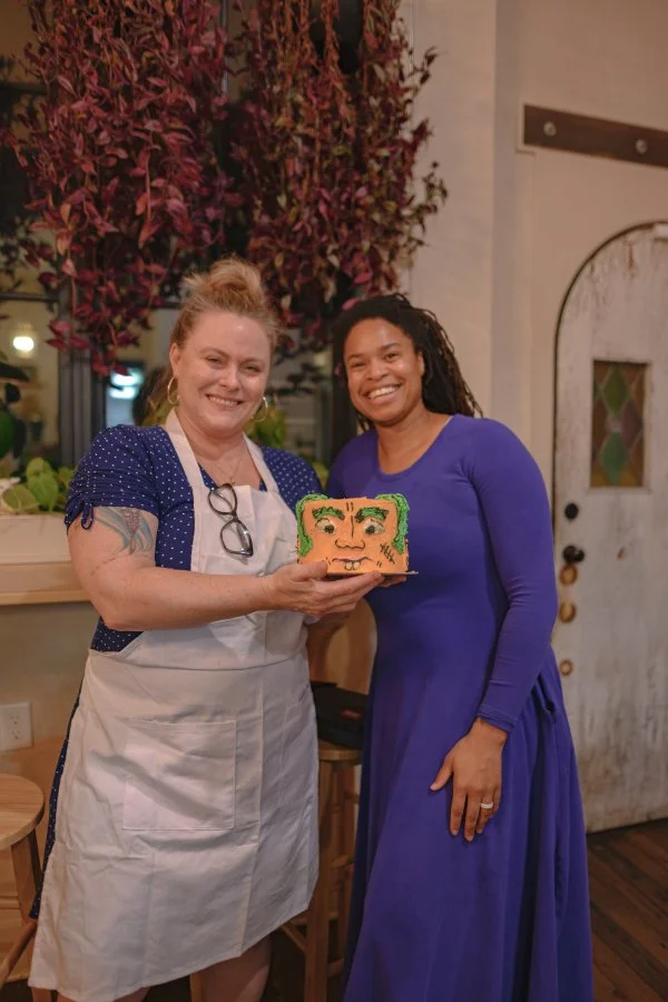 Two women smiling, one in an apron holding a cake with a face design, and the other wearing a blue dress, inside a warmly lit room with purple foliage decor.