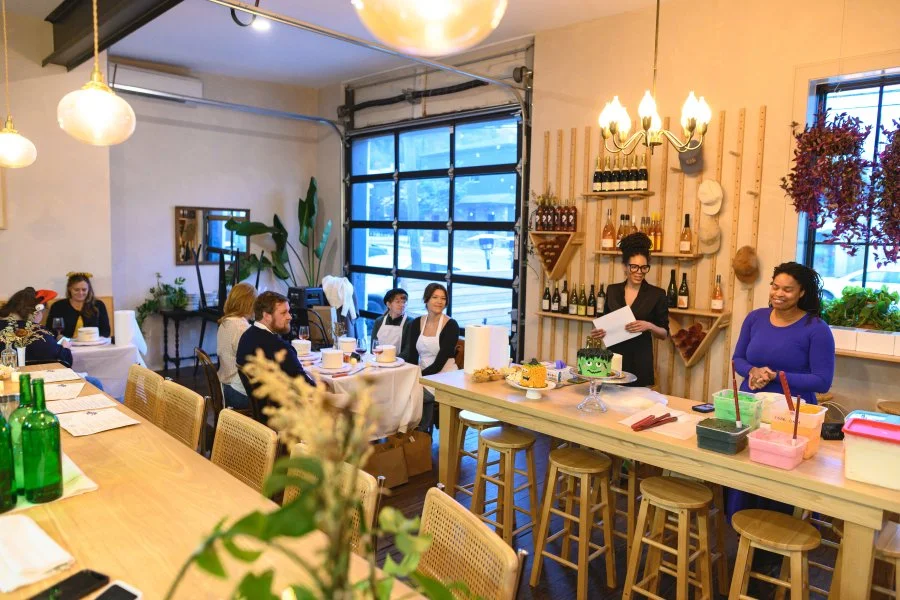 A group of people gathered in a cozy restaurant with a modern interior. Two women are standing behind a kitchen island; one is reading a paper, the other is smiling. Customers are seated at tables, some engaged in conversation. The restaurant is deco