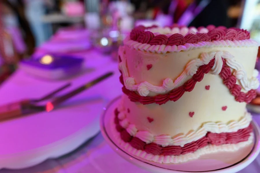 Two-tiered cake decorated with pink and white frosting, small red hearts, and ribbon-like icing patterns, on a white plate at a party or celebration.