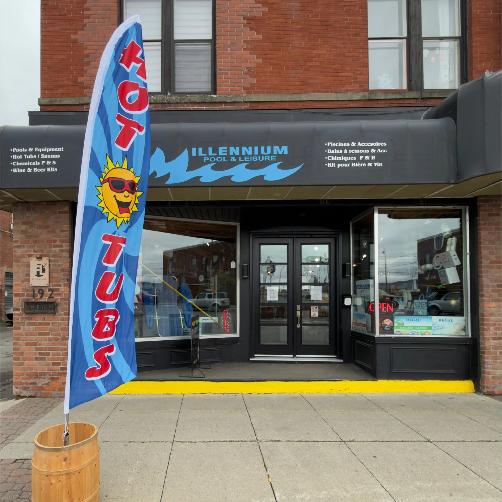 storefront of a pool and leisure shop with a large blue flag with a smiling sun wearing sunglasses, a black awning with the shop's name 'Illennium Pool & Leisure,' and a glass door with an 'Open' sign, along with a yellow-painted curb in front.