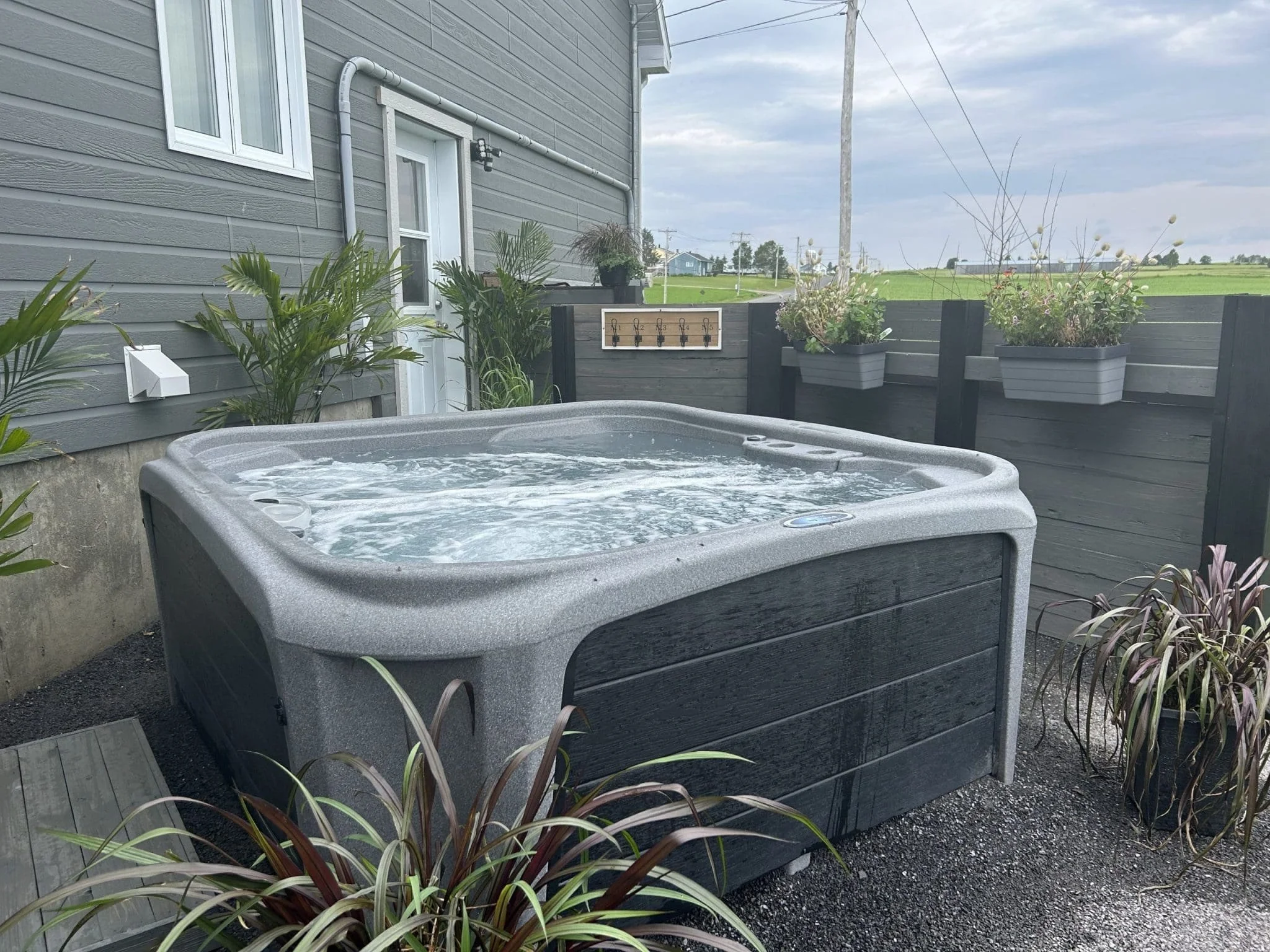 A hot tub on a patio next to a house with gray siding, decorated with potted plants and framed decor on a backyard patio with a rural landscape in the background.