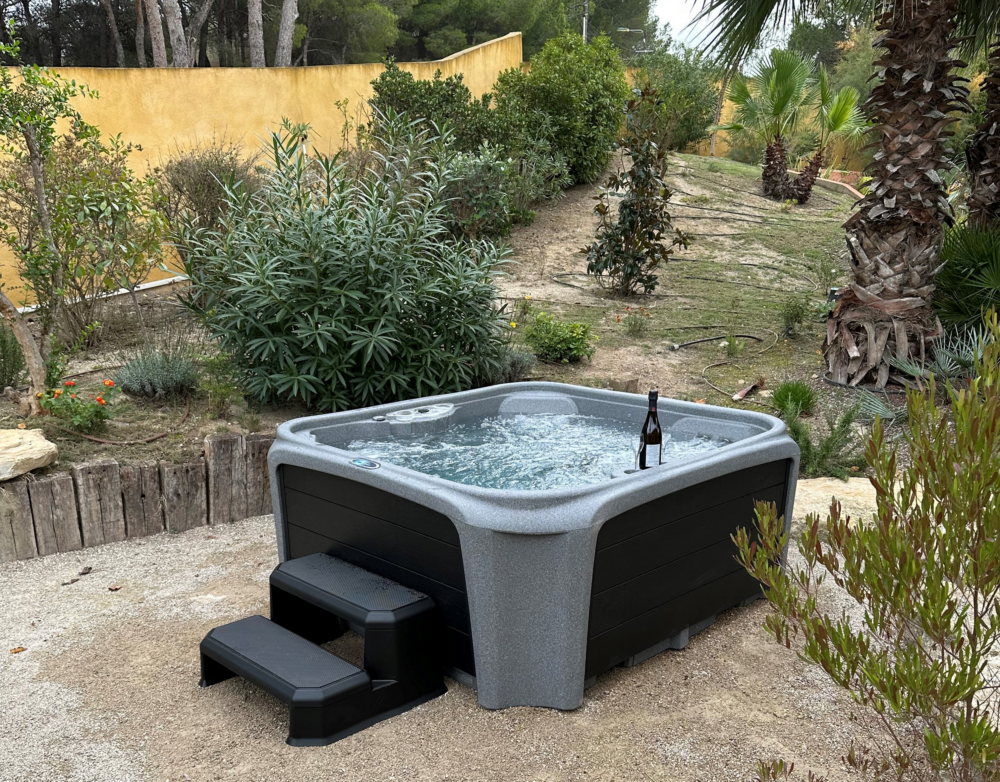 A hot tub with bubbling water and a bottle of wine on the edge is situated in a landscaped backyard with various green plants, bushes, and trees, and a yellow wall in the background.