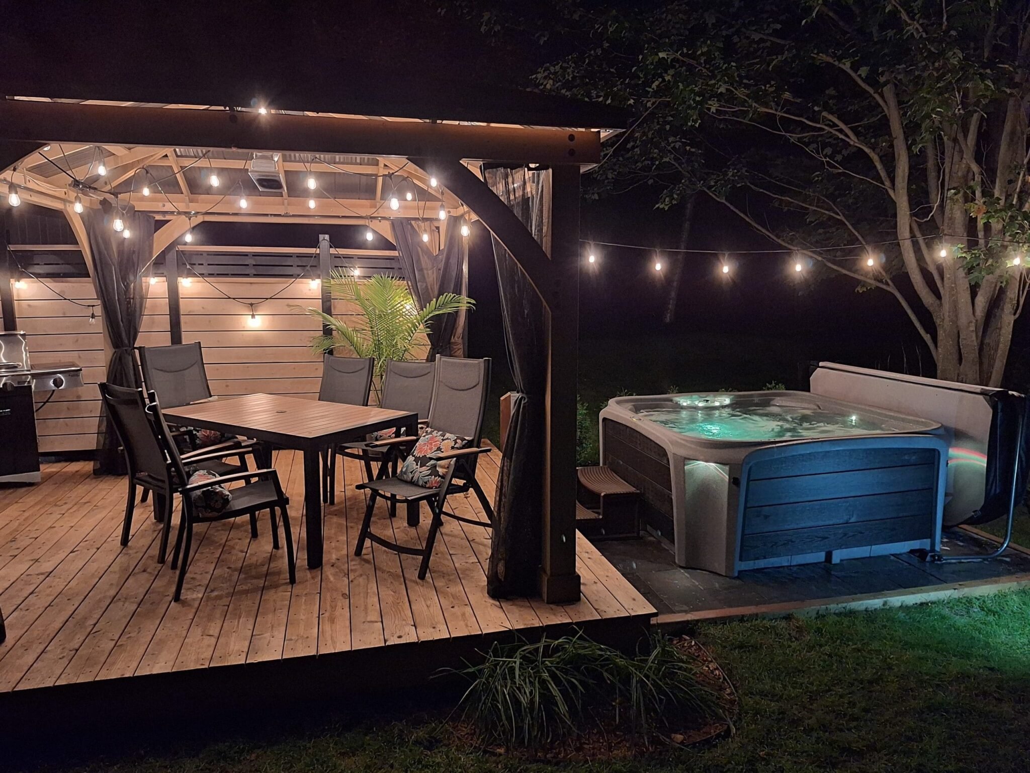 Night view of an outdoor wooden deck with a dining table, chairs, string lights, and a hot tub.