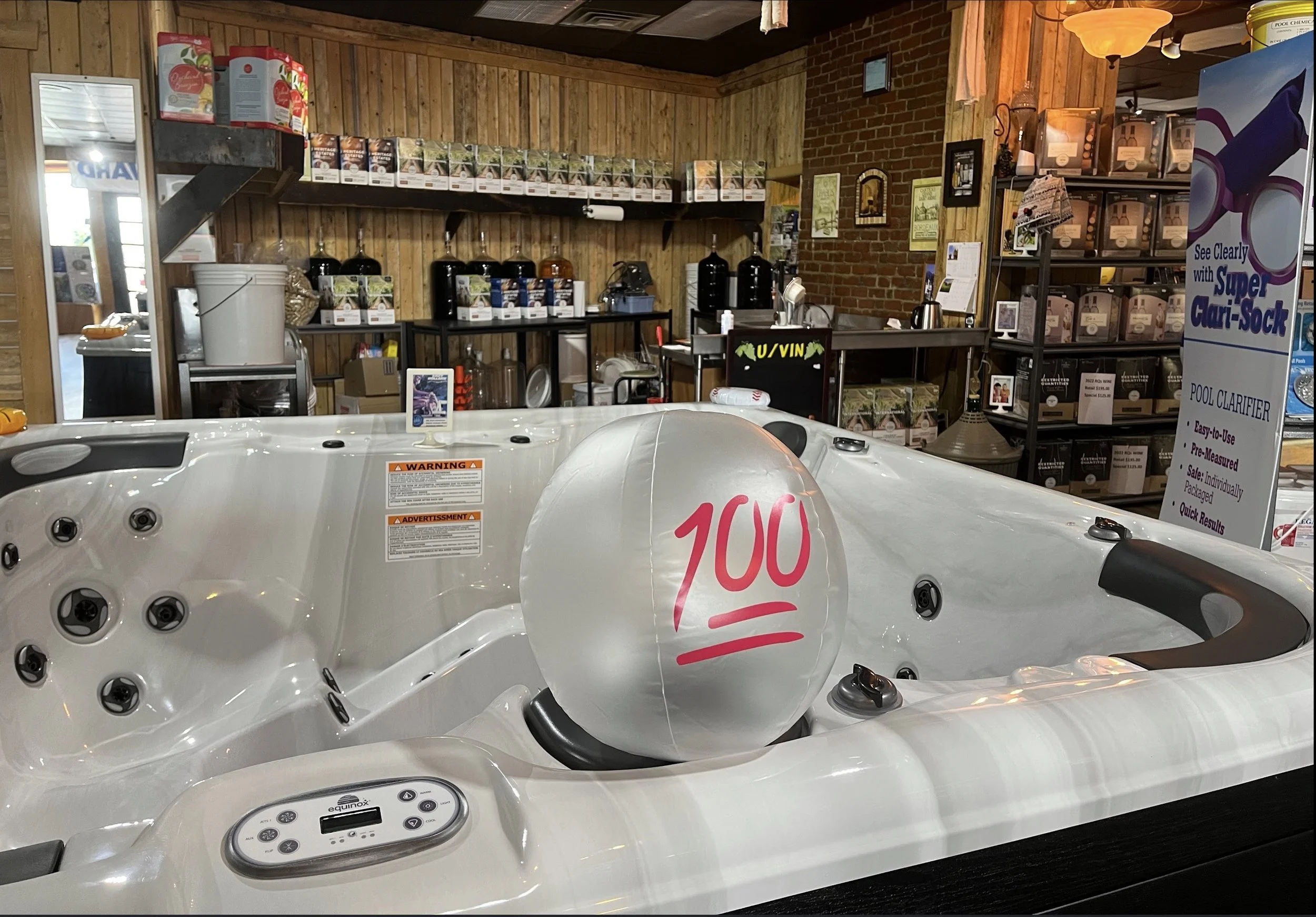 A white hot tub with a large balloon labeled '100' on it, situated inside a wooden and brick room with shelves of supplies and a sign promoting pool clarifier.