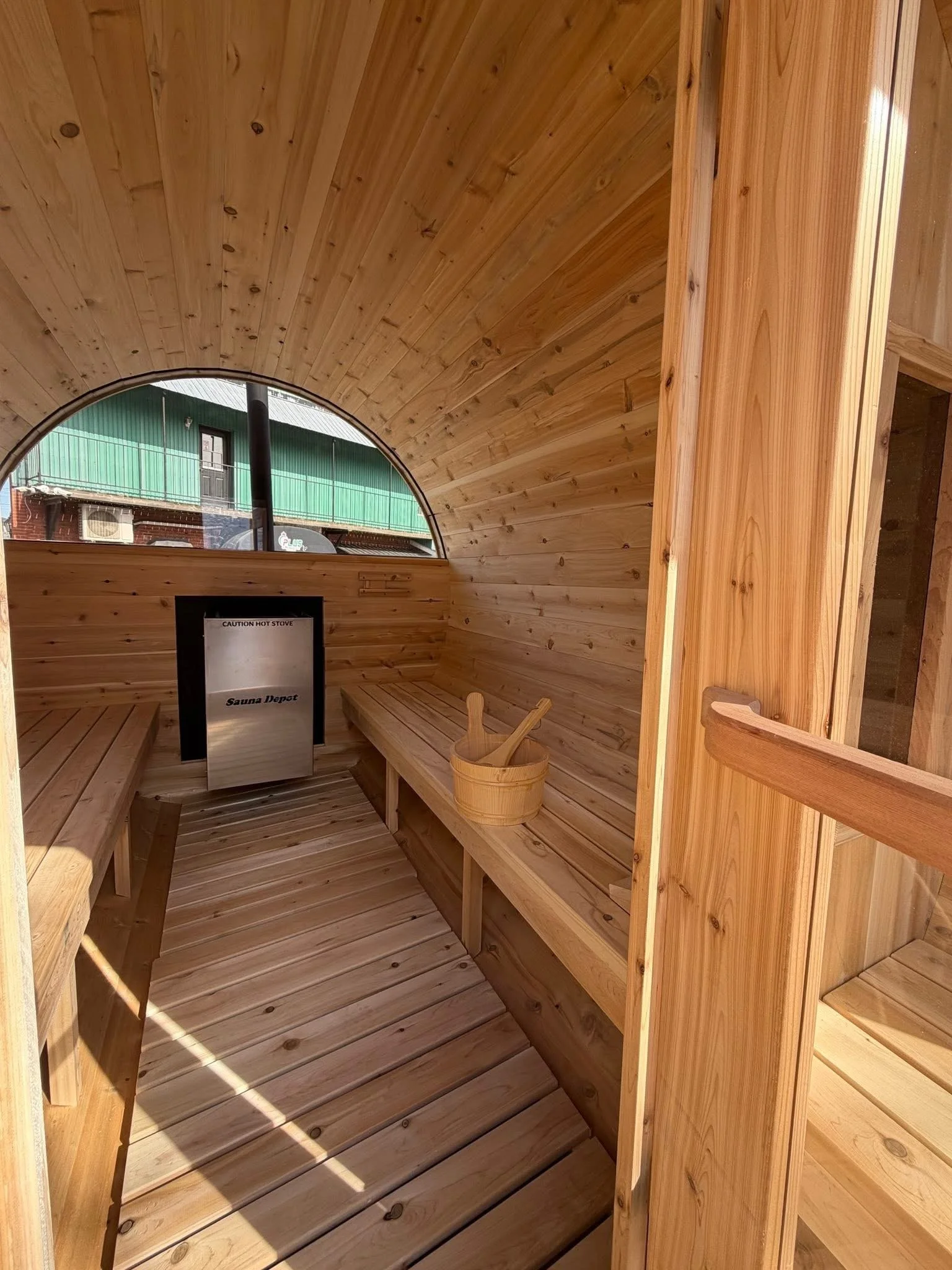 Interior of a small wooden sauna with benches and a bucket with sauna tools, a small window with a view outside.