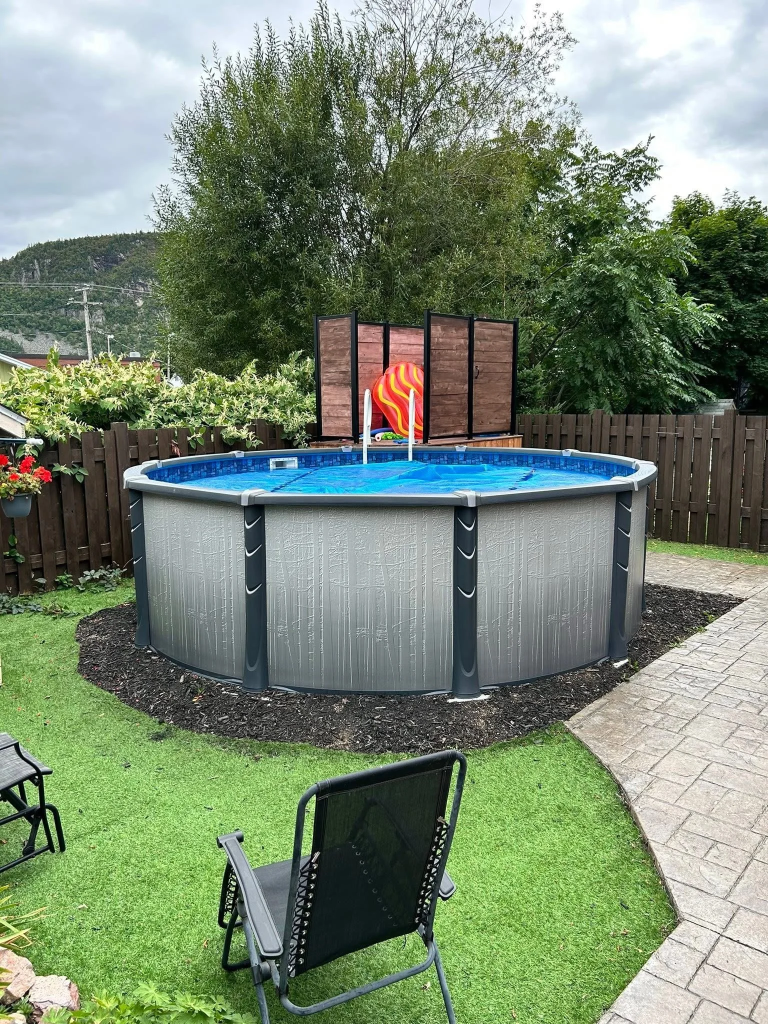 Backyard with above-ground swimming pool on grassy area, surrounded by a wooden fence, with chairs and stone pathway, trees in the background, and pool toys on the pool cover.