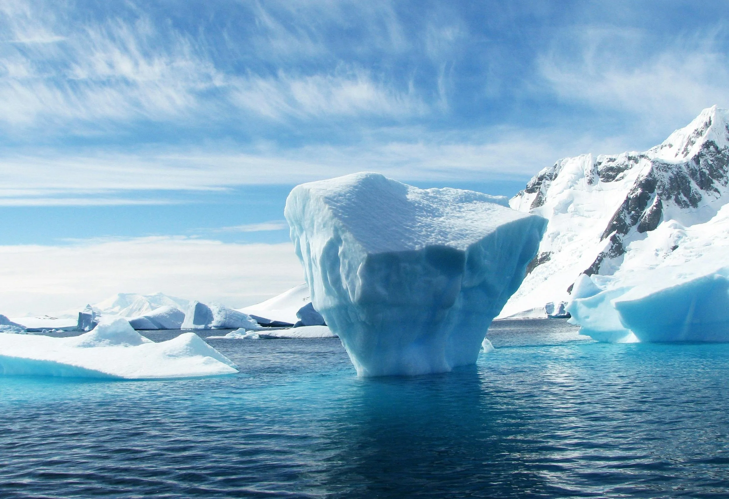 Iceberg floating in the ocean with mountains and a cloudy sky in the background.