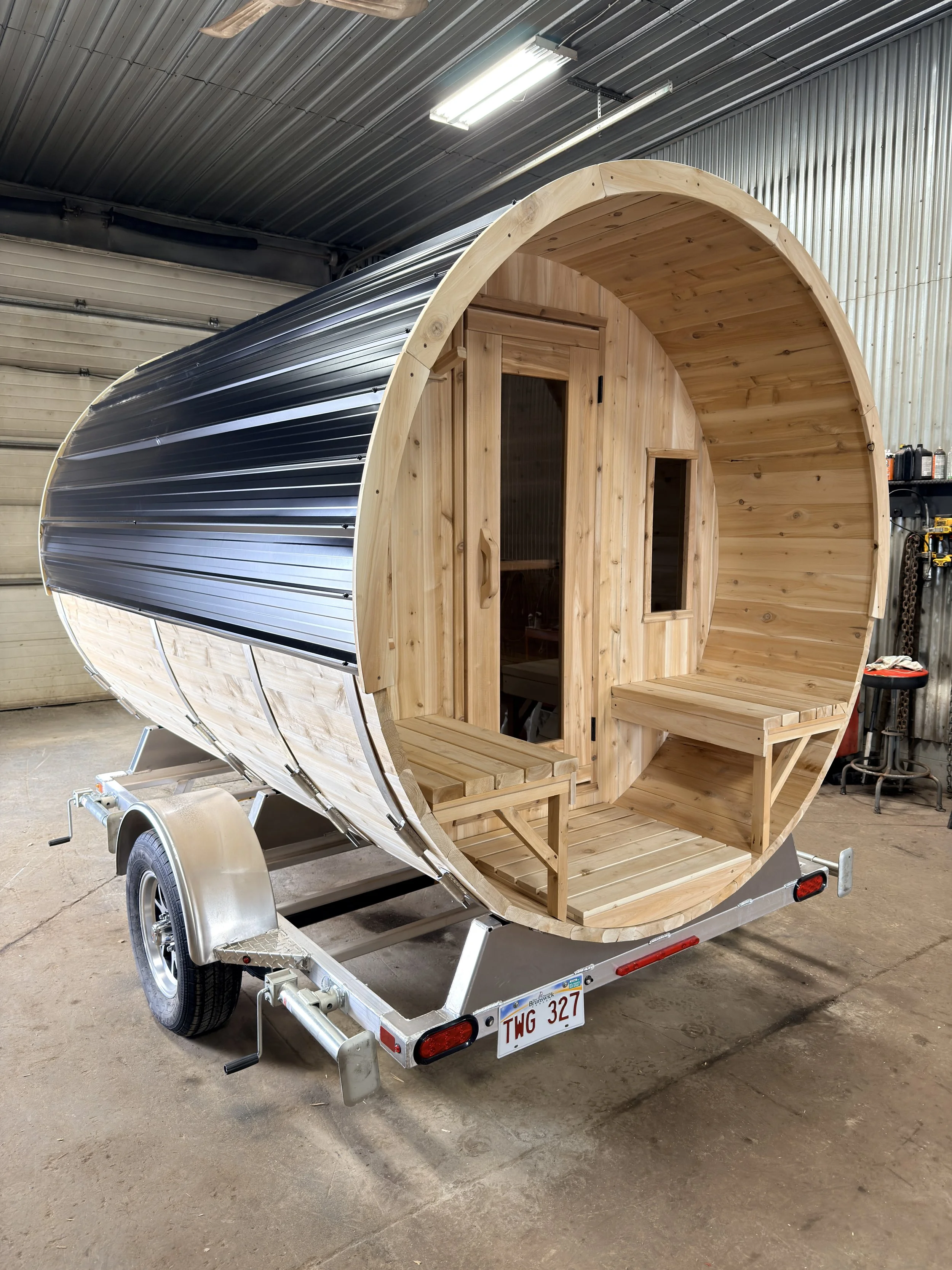 A barrel sauna with a black metal exterior roof and a wooden interior designed like a tiny house, mounted on a trailer with a single axle, inside a garage.