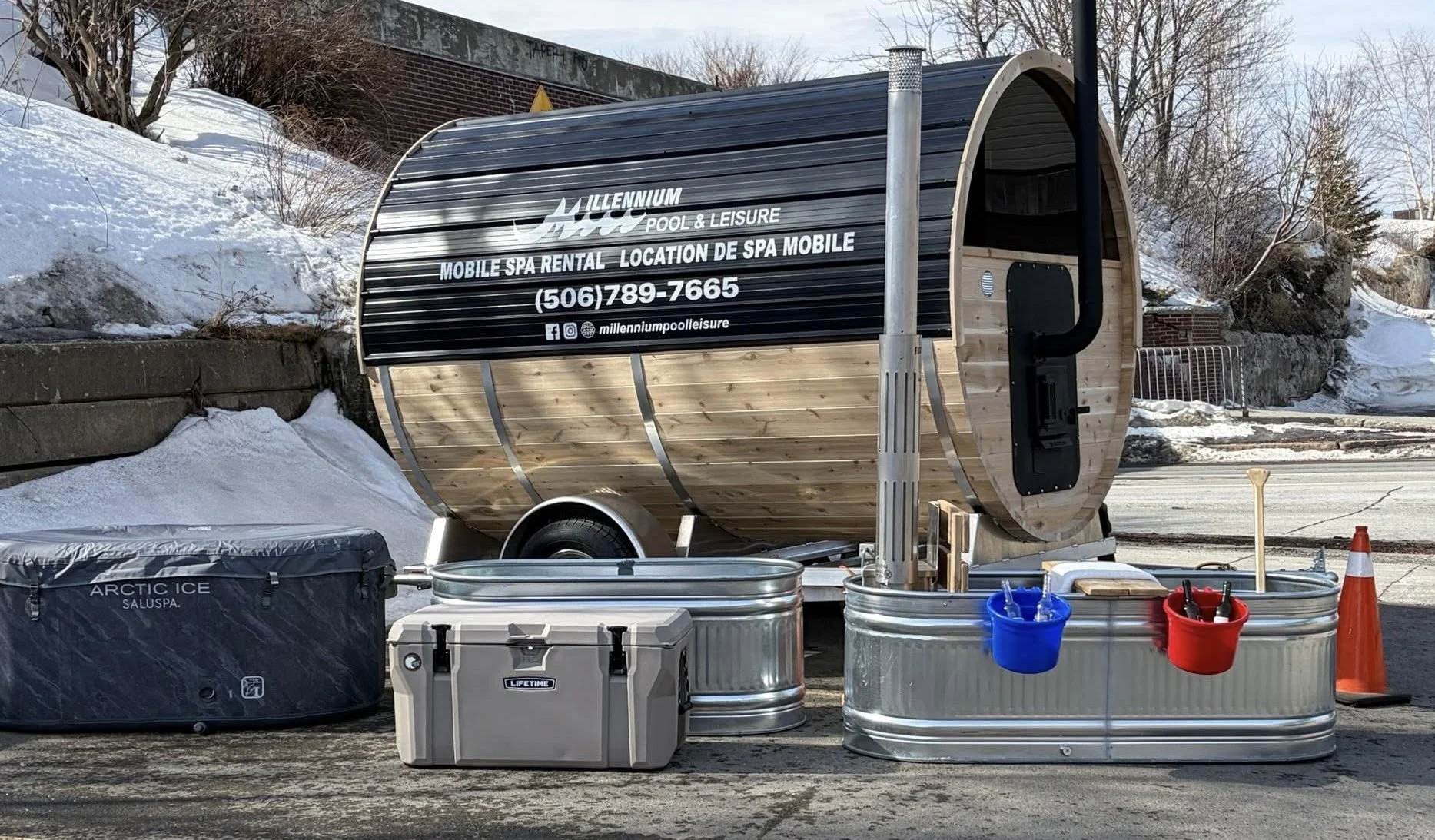 A mobile spa trailer with containers and equipment, parked outdoors on a winter day, with snow on the ground and trees in the background.