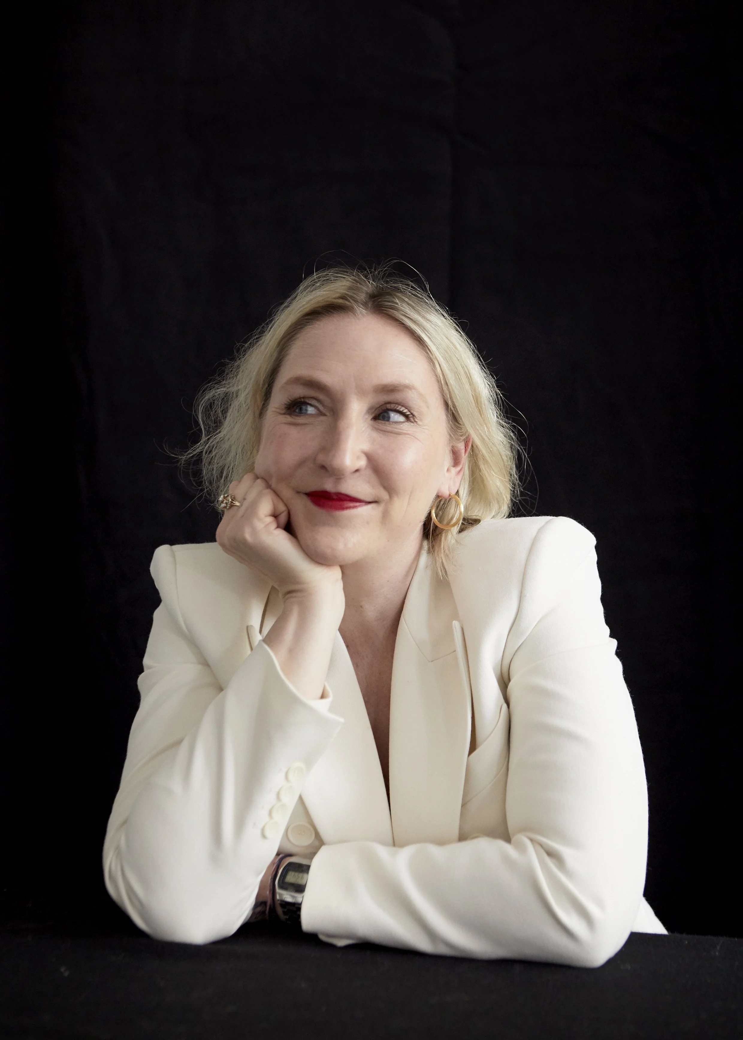 A woman with blonde hair, wearing a white blazer, resting her chin on her hand, smiling slightly, with a dark background.
