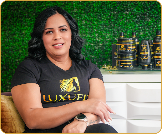 Woman with dark hair and a black t-shirt sitting in front of a green leafy background and hair care products.