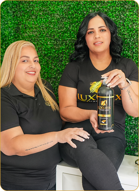 Two women in black LUXFIX uniforms are posing in front of a green wall, with one woman holding a bottle of shampoo.