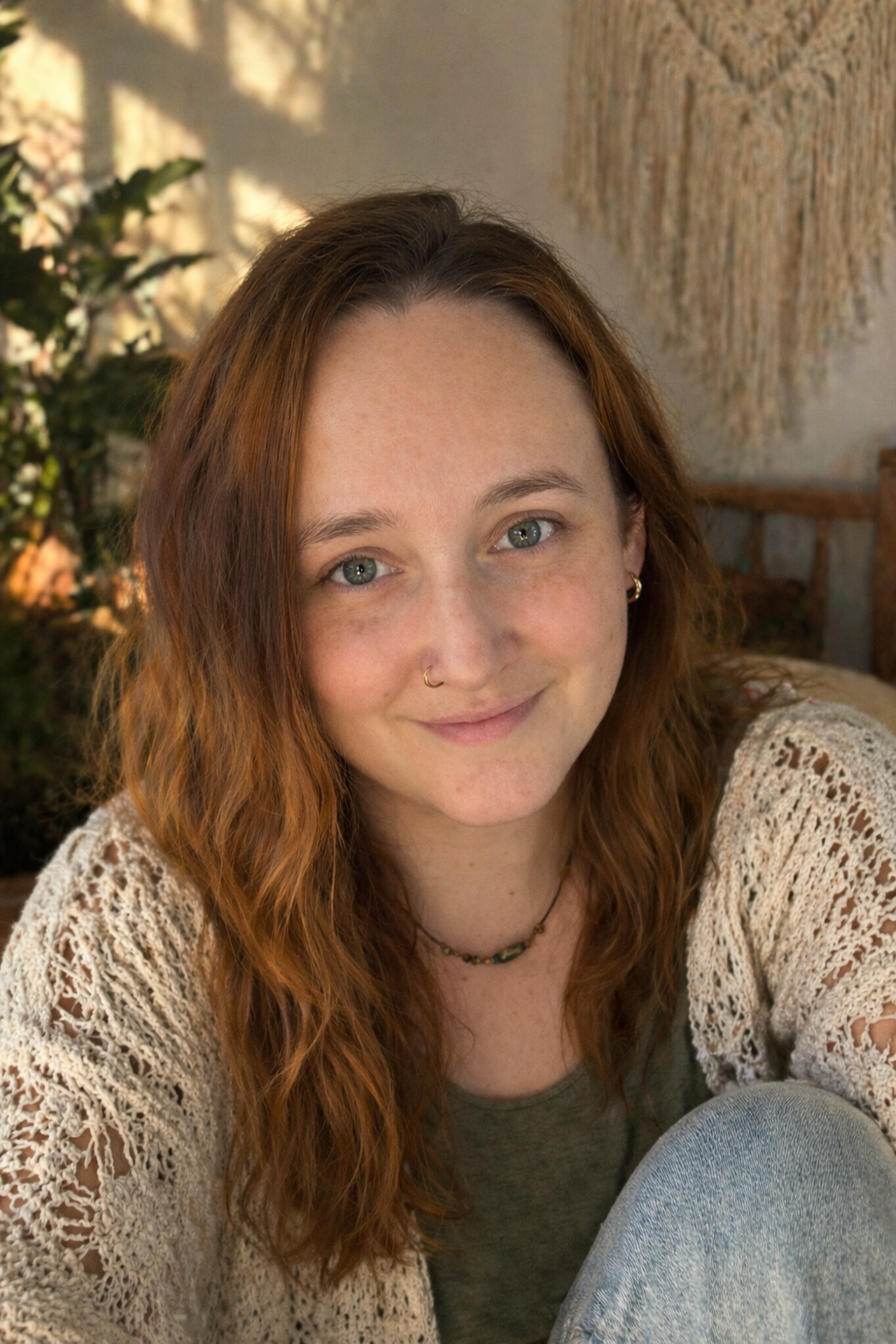 A young woman with wavy red hair, light skin, and blue eyes smiling at the camera, wearing a beige crochet cardigan over a green top, with a necklace and earrings, indoors with plants and a wall hanging behind her.
