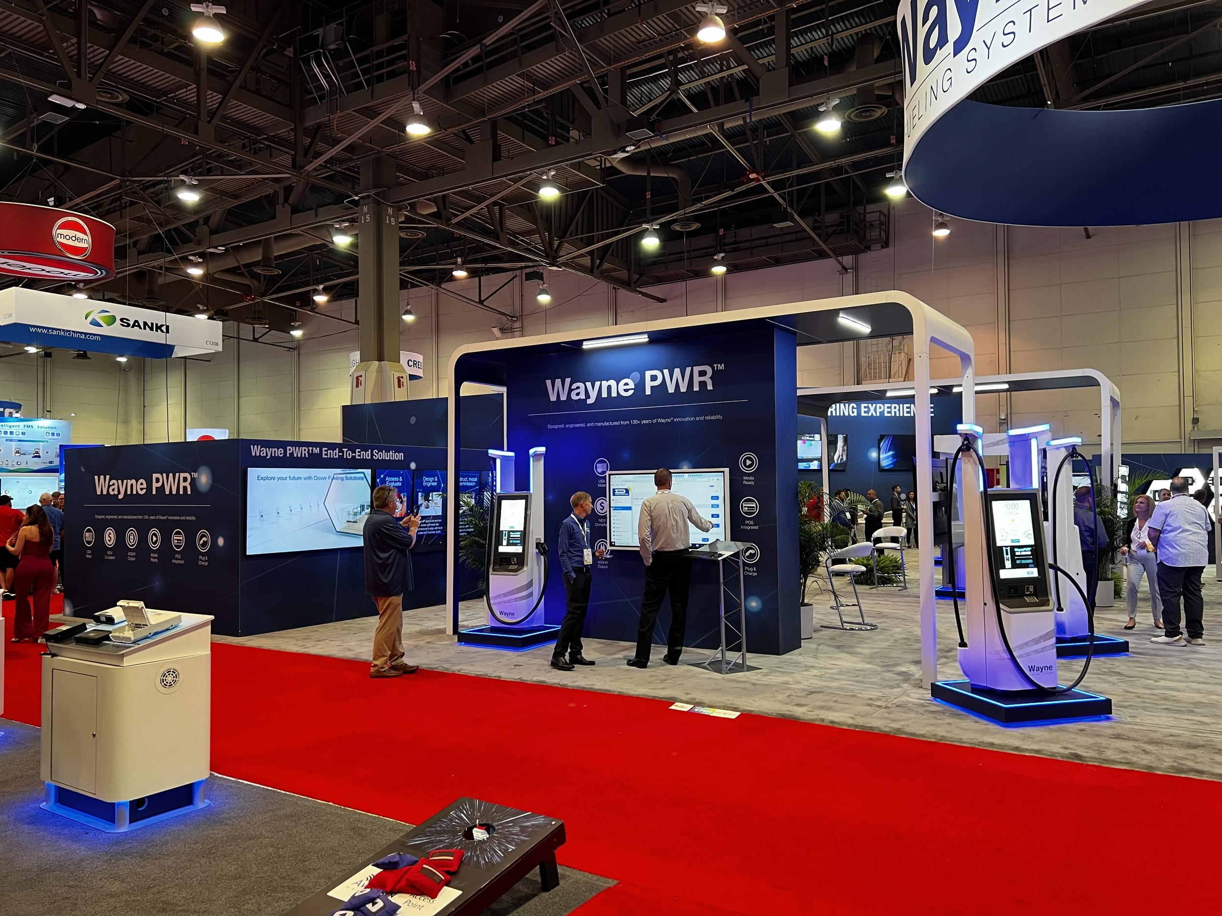 Trade show booth displaying Wayne PWR electric vehicle charging stations with signage and people interacting, in a large exhibition hall with high ceilings and overhead lighting.