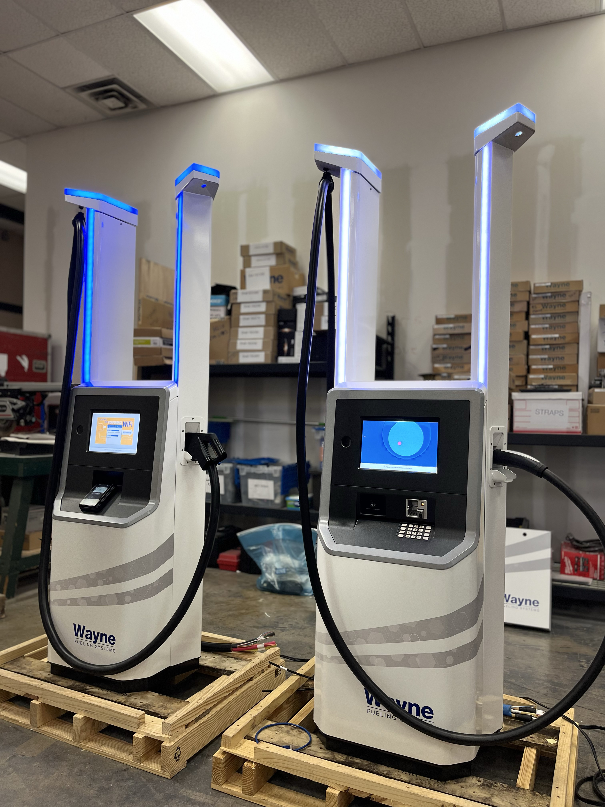 Two Wayne fueling systems with digital screens and fuel nozzles, positioned on wooden pallets in an industrial setting with shelves and boxes in the background.
