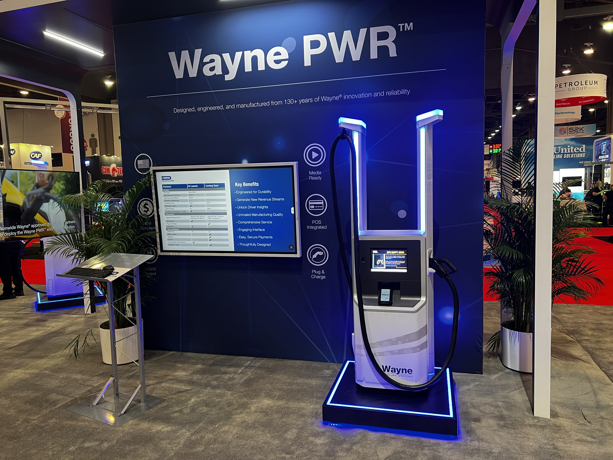 Wayne PWR electric vehicle charging station with digital display screen, plant decor, and informational signage at an exhibition.