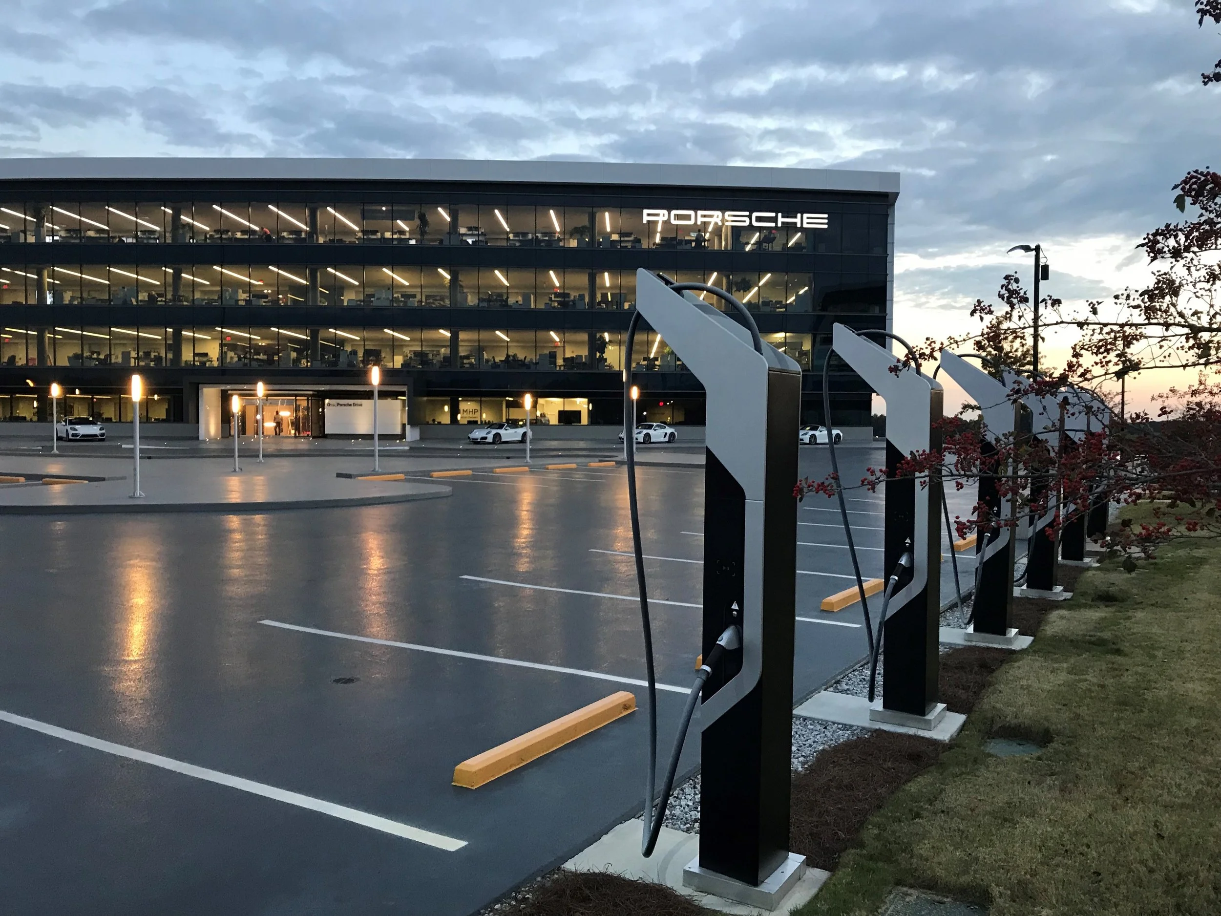View of a Porsche dealership building with charging stations in the parking lot during dusk, and several cars parked near the entrance.