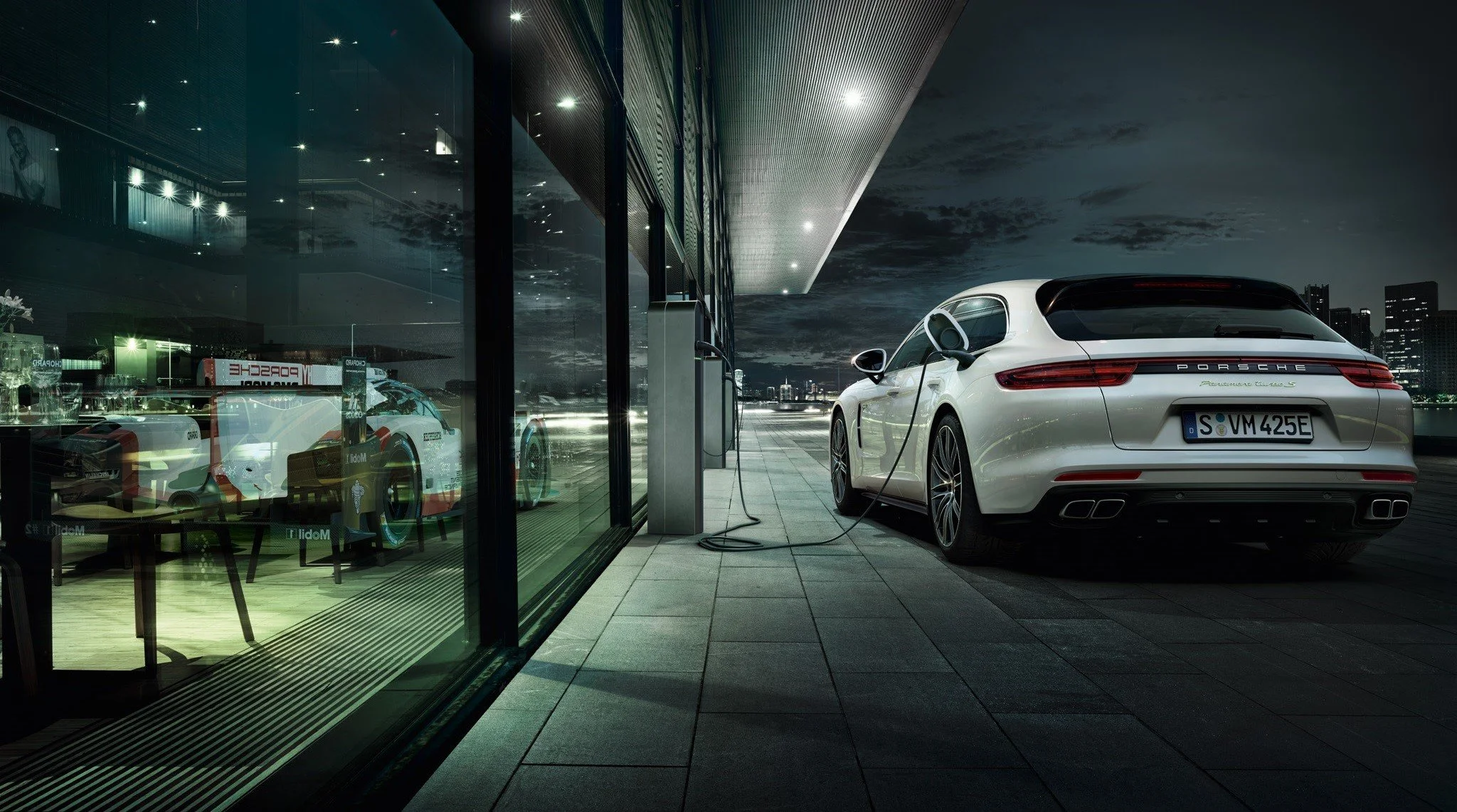 A white Porsche sports car connected to an electric charging station outside a modern building at night, with reflections of the car and city skyline in the glass window.