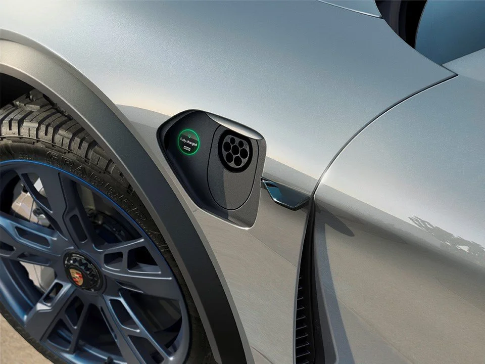 Close-up of an electric vehicle charging port on a silver car with a Porsche emblem on the wheel.
