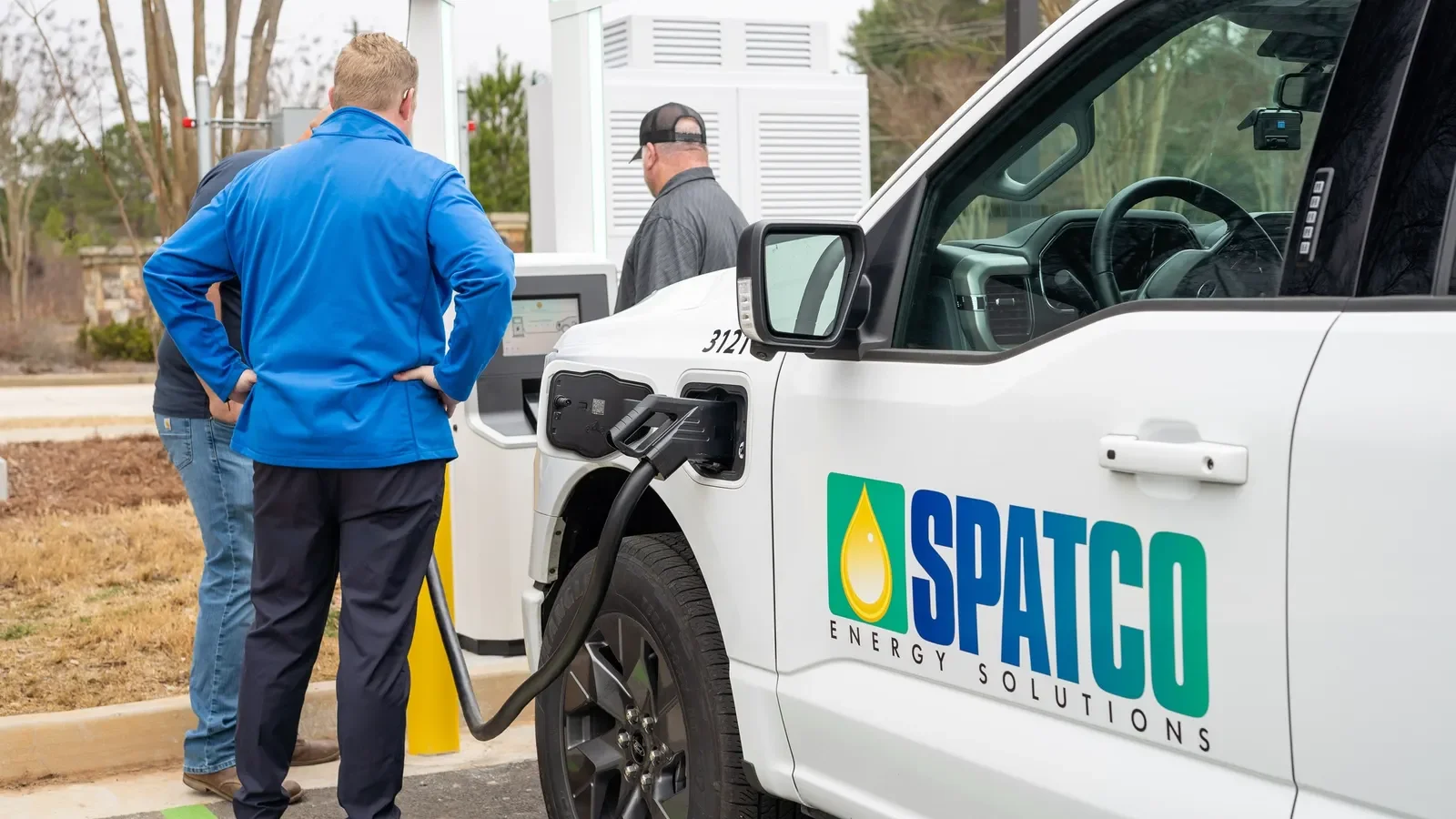 Two men charging an electric vehicle at a SPATCO energy solutions station, with a white electric truck parked at the charger.