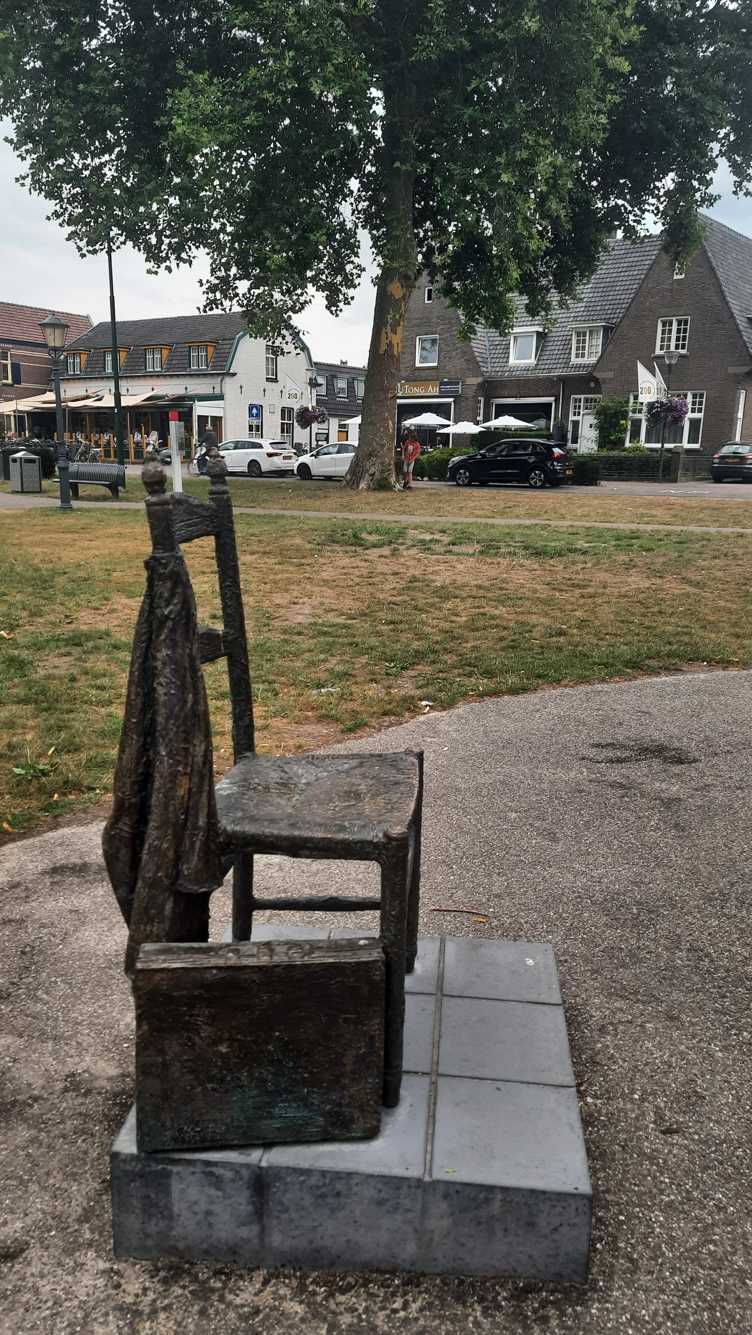 A metal sculpture of a chair with a coat hanging on its backrest, mounted on a concrete pedestal, outside on a sidewalk in a town square. ( that is fro, Van Gogh's native village Nuenan )