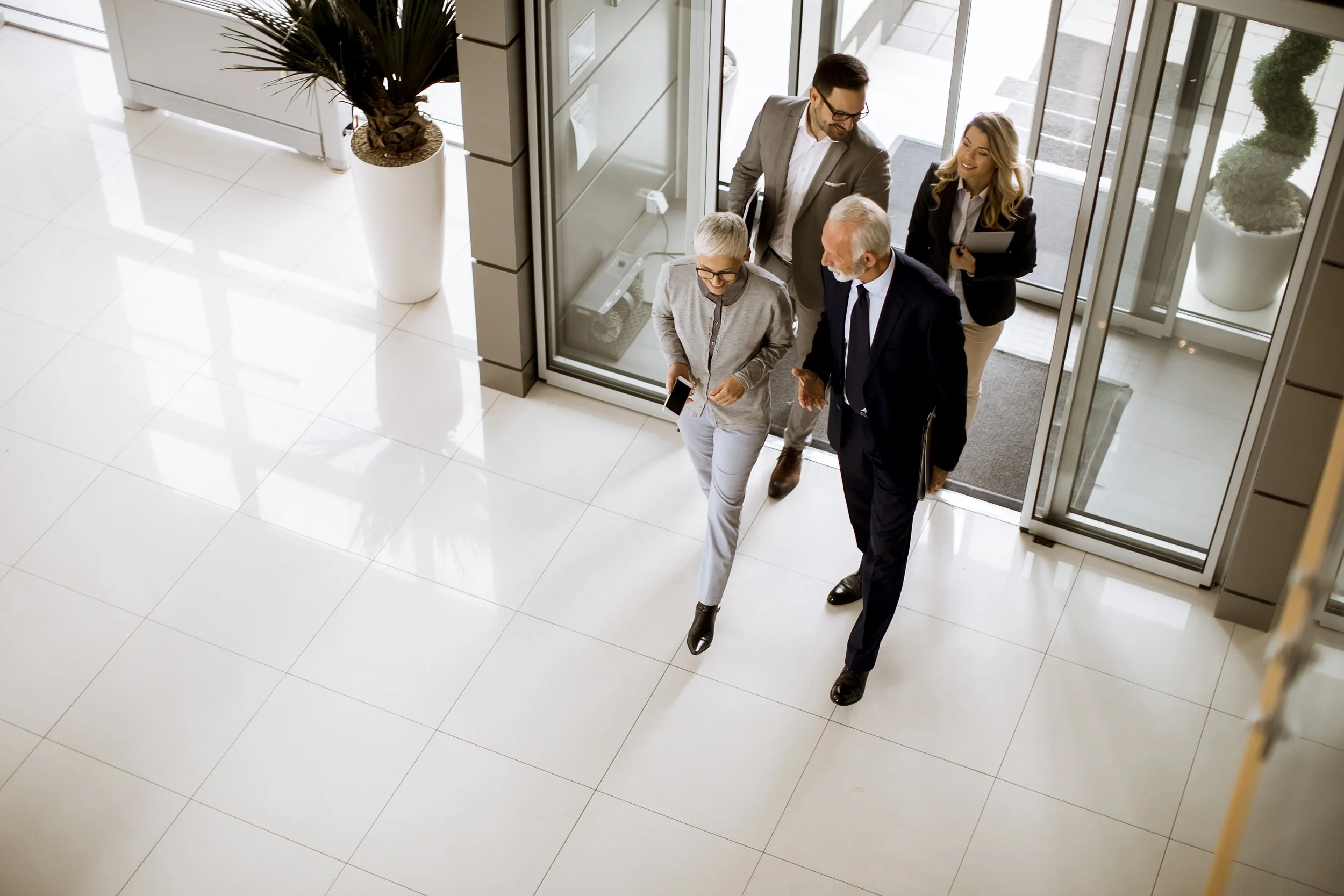 Four people entering a modern building through glass doors, with two women and two men dressed in business attire, one of the women holding a tablet and the other holding a smartphone.