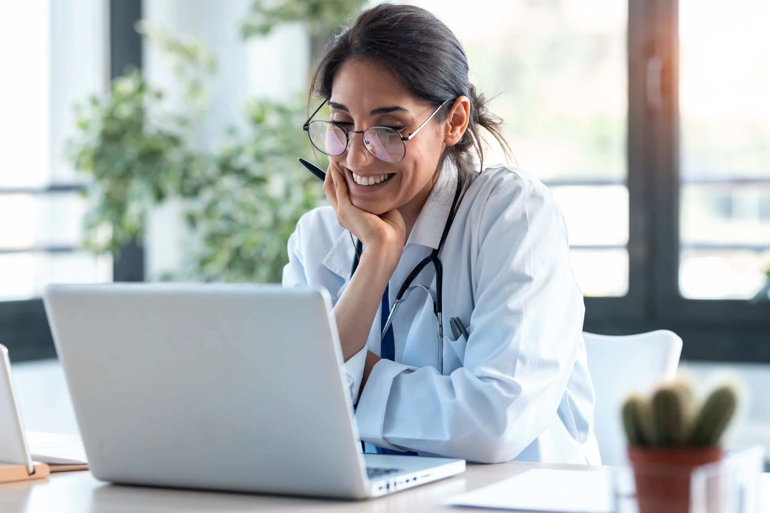A female doctor wearing glasses, with a stethoscope around her neck, smiling while sitting at a desk with a laptop. The background has large windows and plants.