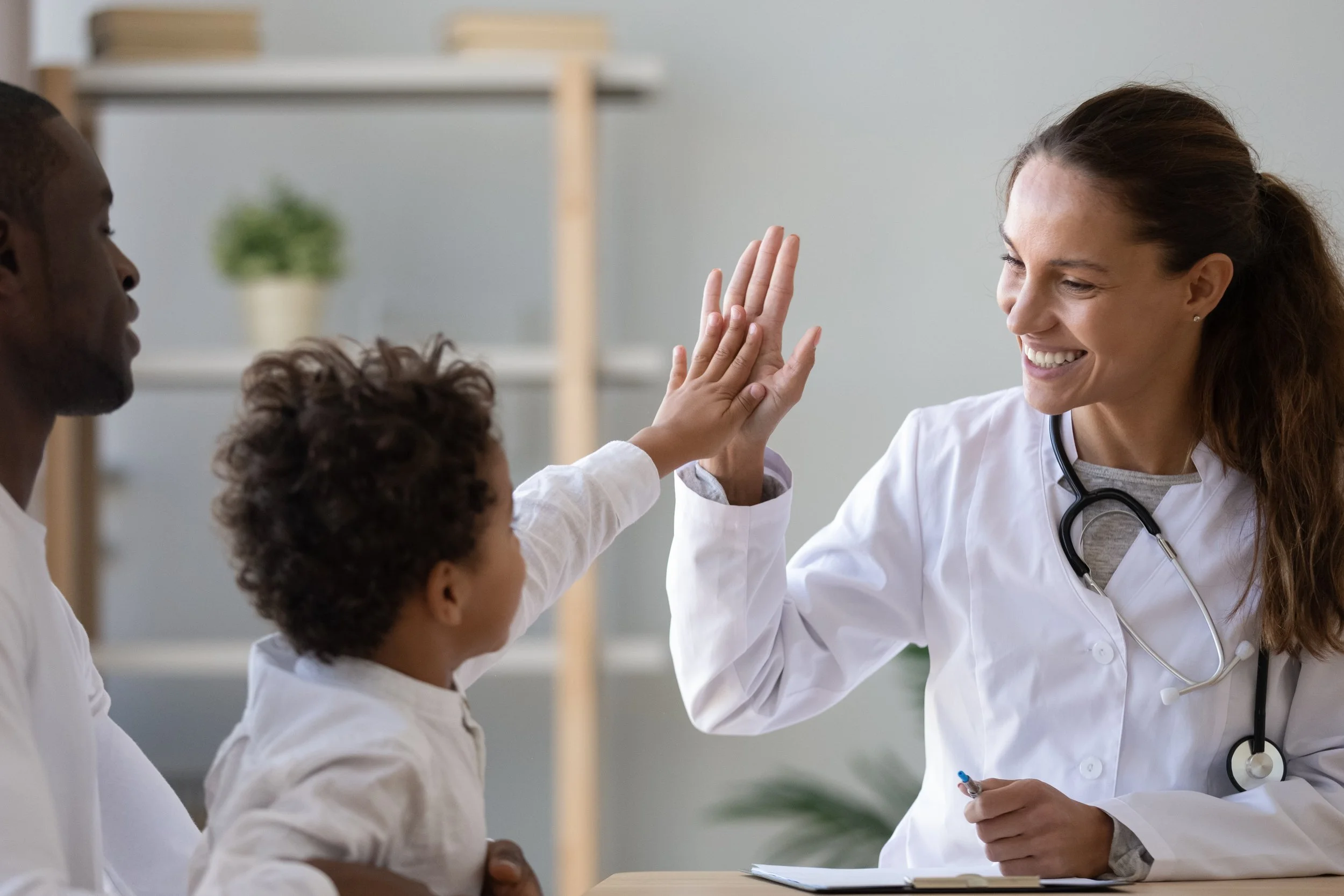 A female doctor smiling and giving a high five to a young patient, with a man and a child in the background, all wearing white medical coats in a clinical setting.