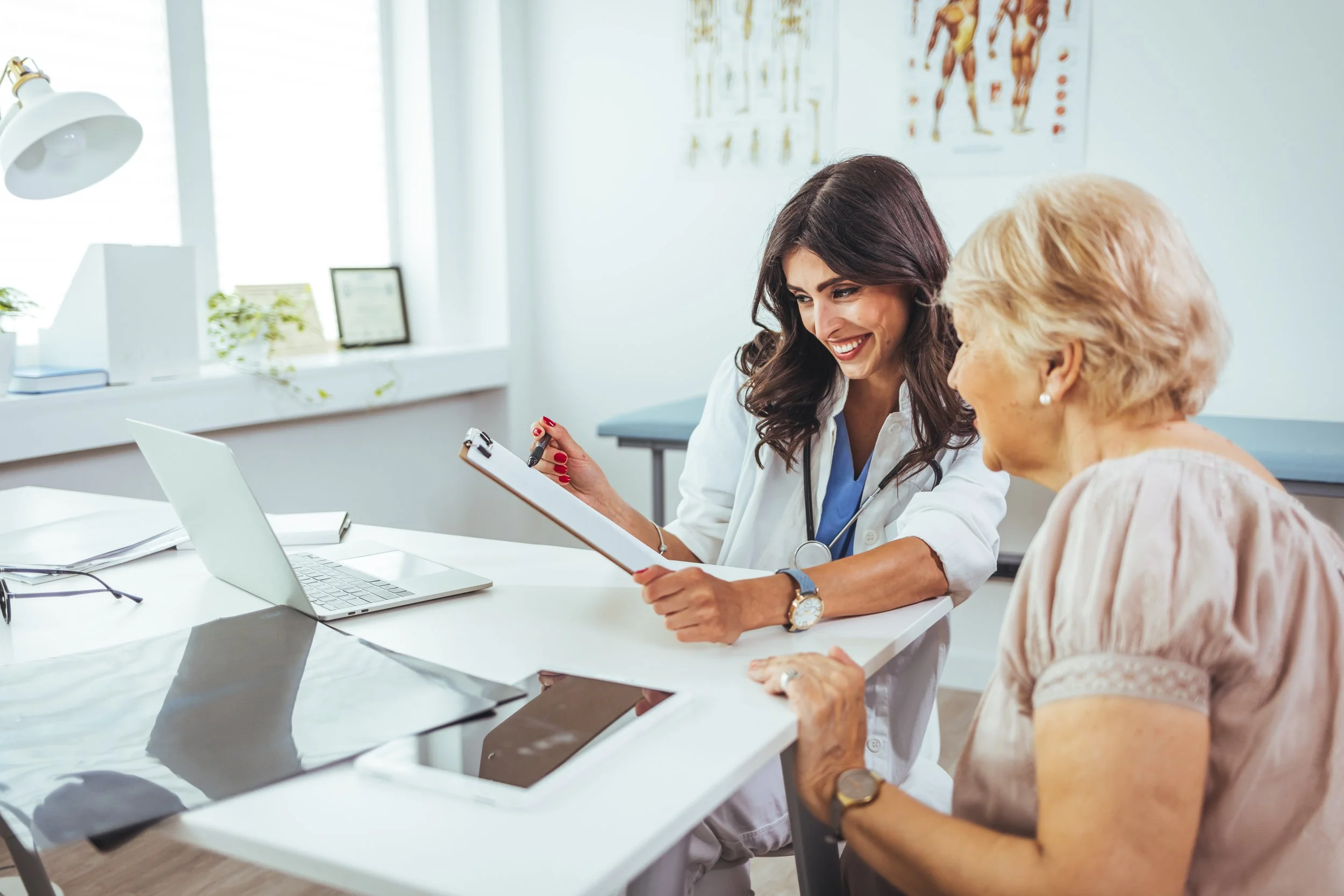 A female healthcare professional in a white coat and stethoscope discussing with an elderly woman at a desk in a medical office. The healthcare professional is smiling and holding a clipboard, while the elderly woman is listening. There is a laptop and some documents on the desk, with anatomical posters on the wall and a window in the background.