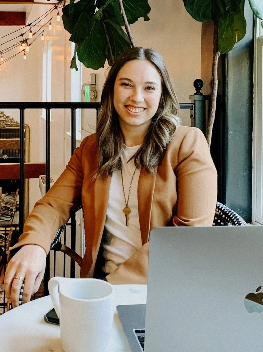 A young woman with wavy brown hair smiling while sitting at a table in a cozy cafe or restaurant. She is wearing a tan blazer over a white top, with a necklace. There are plants, a laptop, a white mug, and decorative string lights in the background.