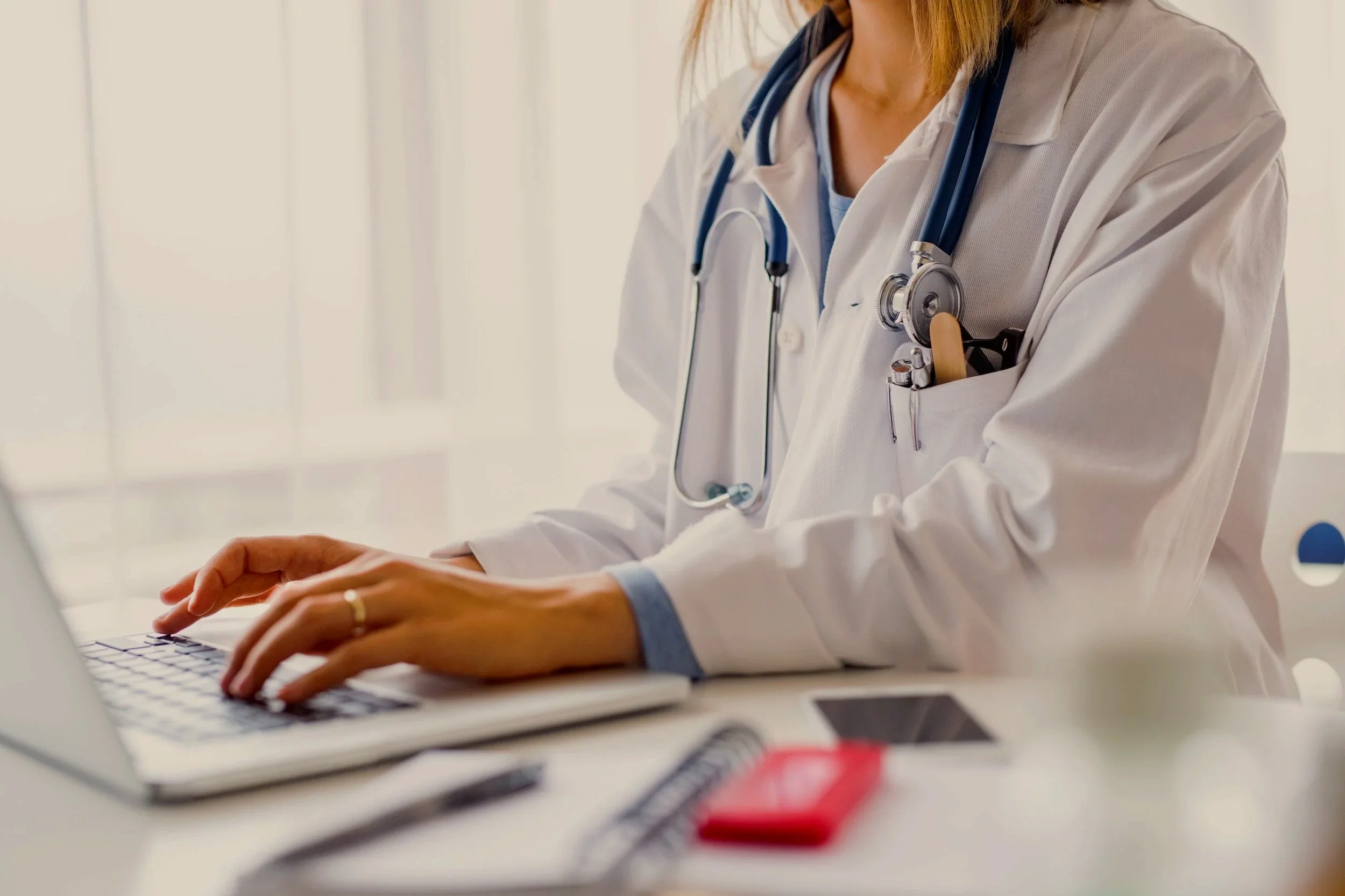 A healthcare professional, wearing a white coat and stethoscope, typing on a laptop in an office or clinic setting.