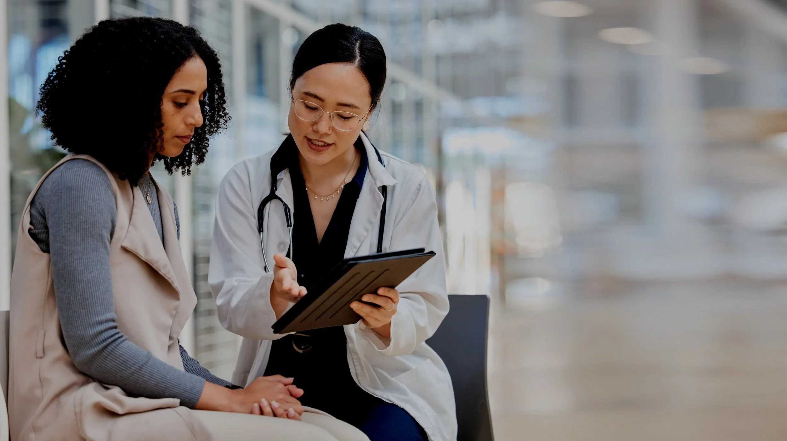 A female doctor consulting with a female patient at a hospital or clinic waiting area, with the doctor showing something on a tablet device.