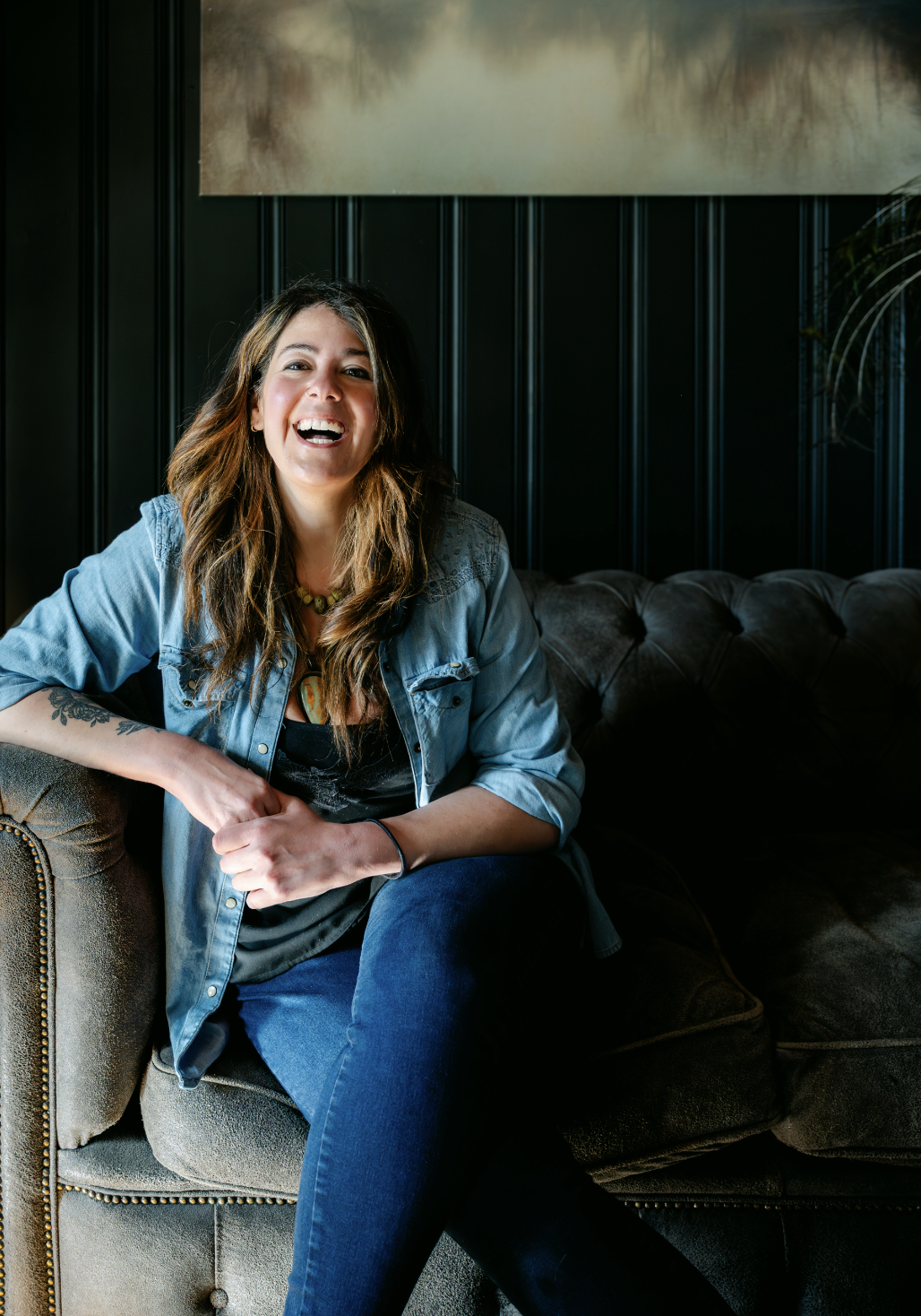 A woman with wavy brown hair wearing a denim jacket sitting on a gray couch, smiling and laughing, with a dark paneled wall behind her.