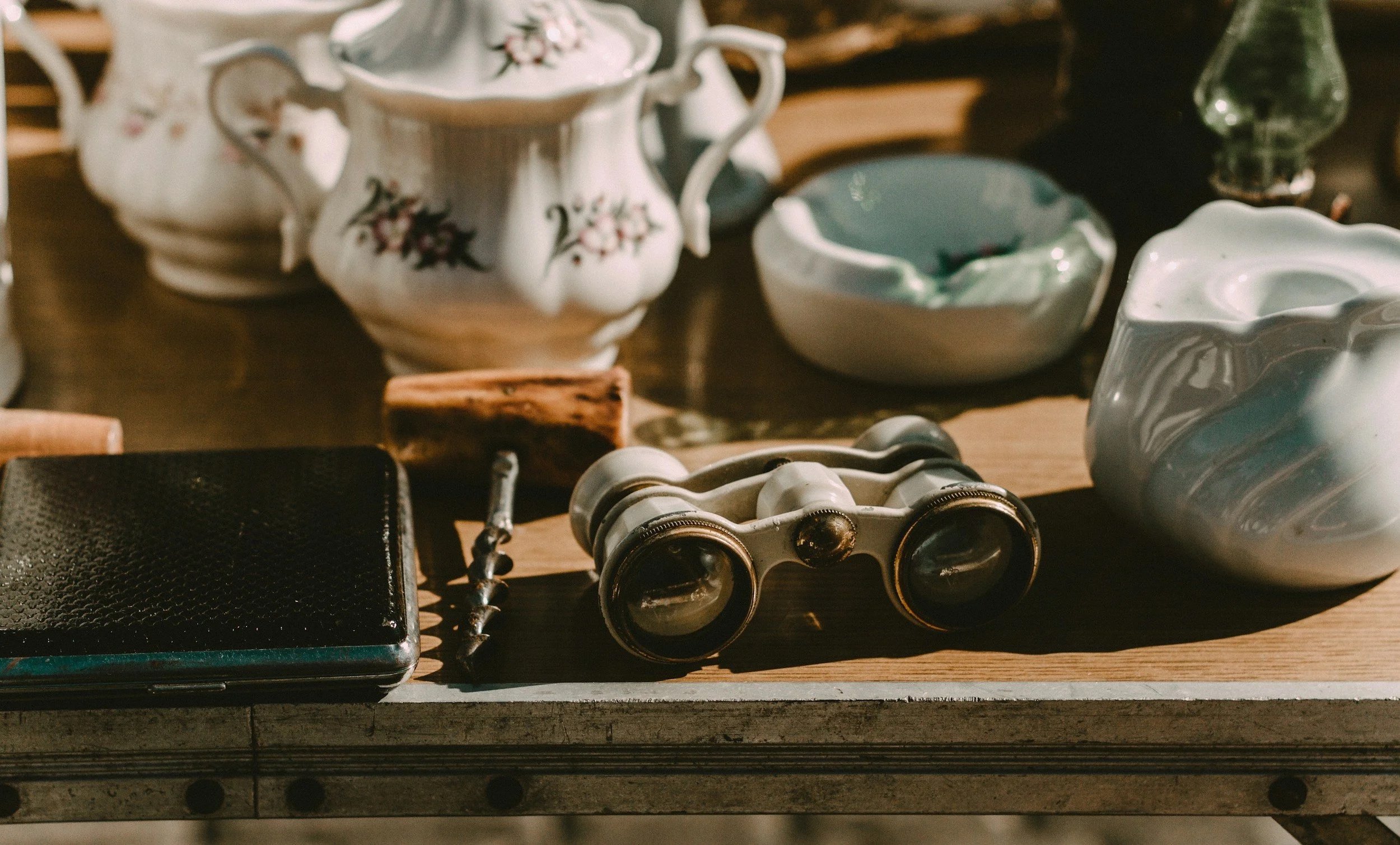 Vintage binoculars, a black wallet, a cork, and ceramic bowls with floral patterns on a wooden table.