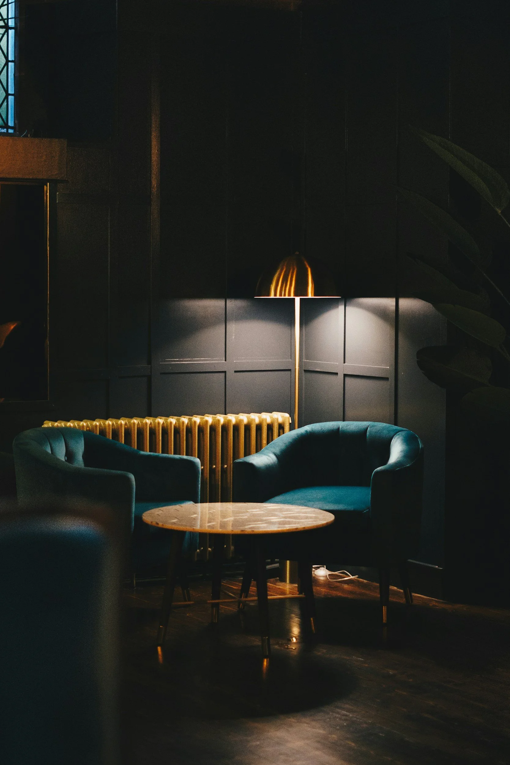 Cozy seating area with two teal armchairs and a small wooden table, illuminated by a brass floor lamp in a dimly lit room with black paneled walls, a radiator, and a large plant.