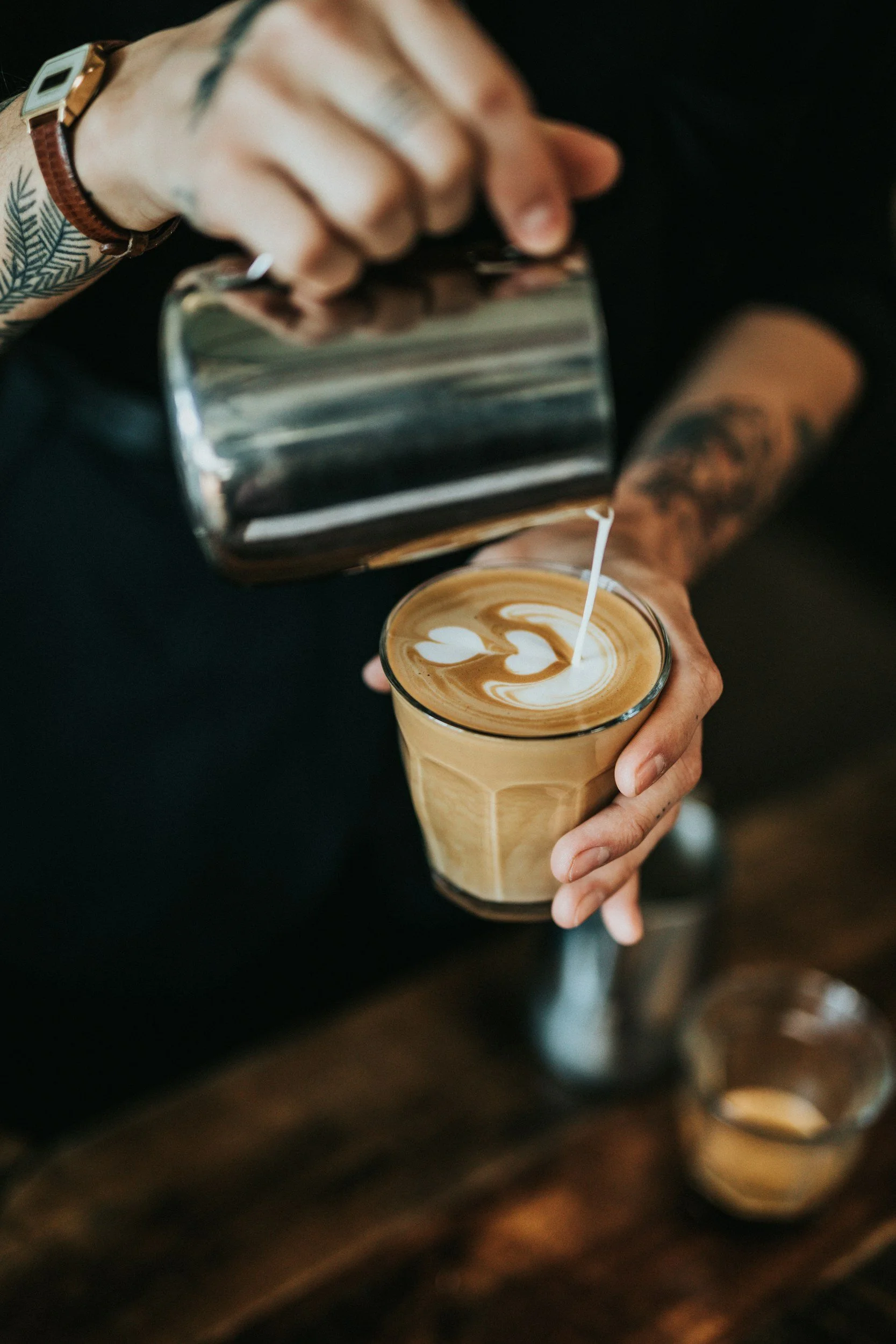 A person pouring steamed milk into a glass of coffee, creating latte art with heart and leaf shapes.