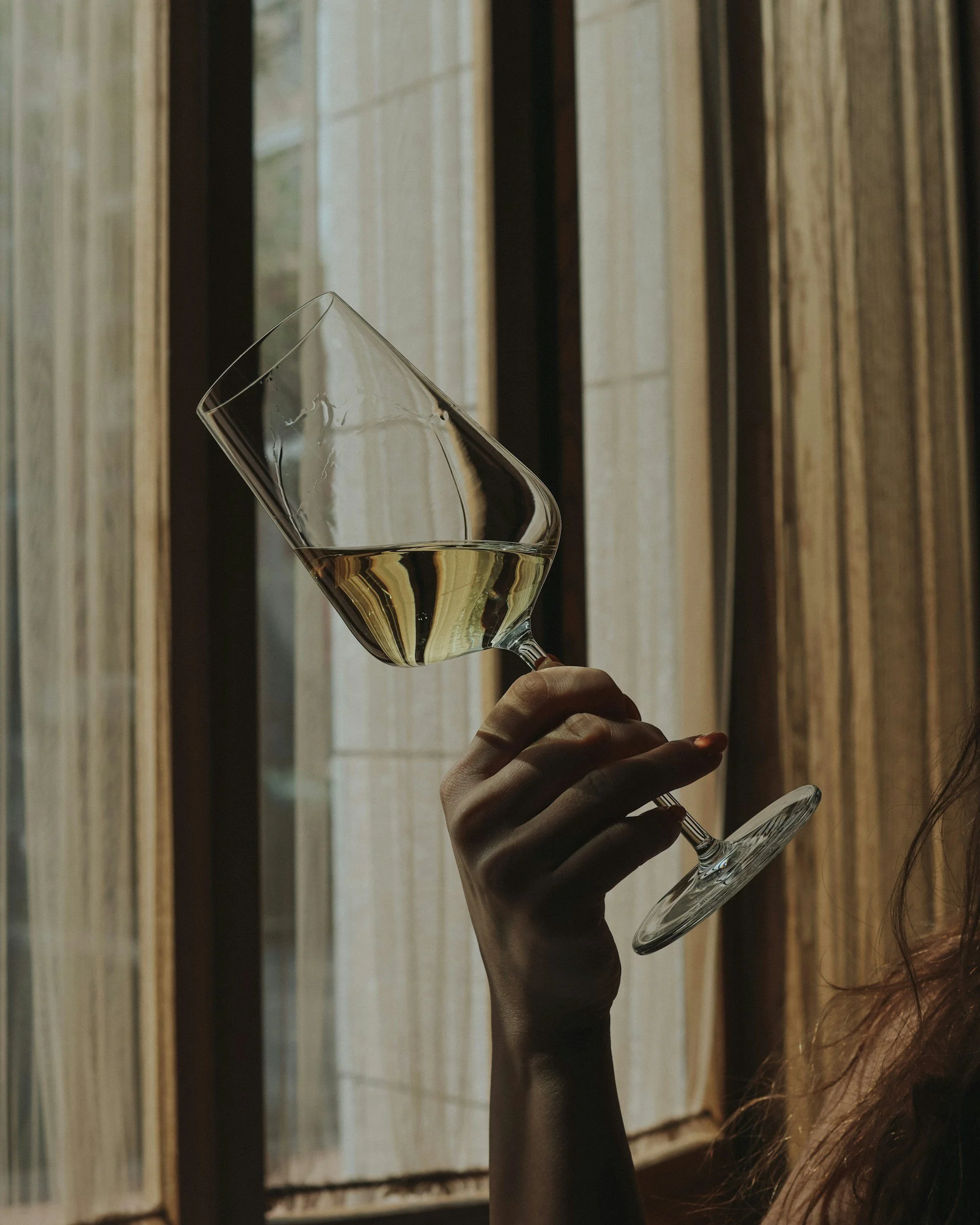Close-up of a hand holding a tilted glass of white wine in front of a window with curtains.