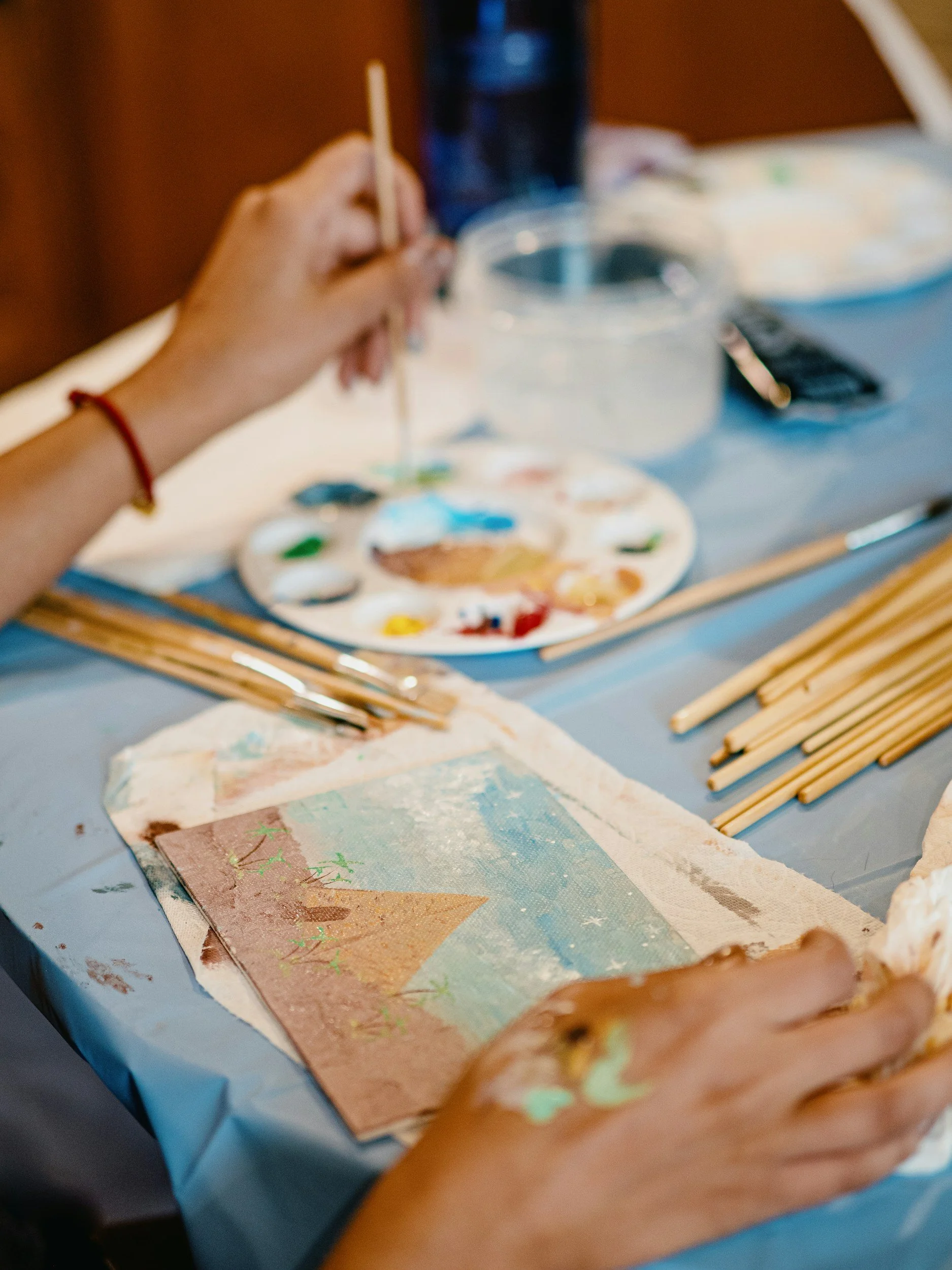 Person painting a landscape on canvas with a palette of paint nearby and wooden skewers on the table.