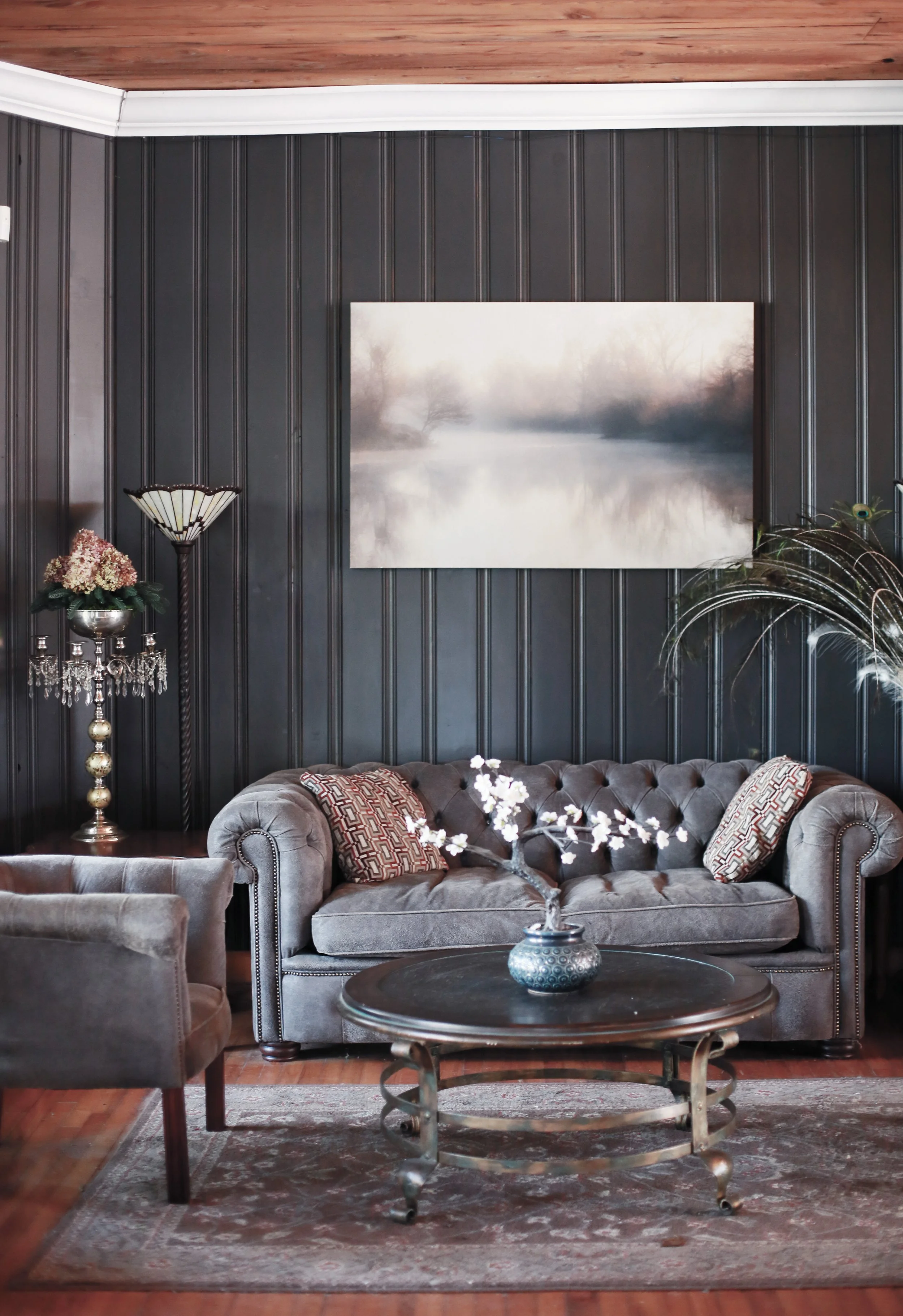 Living room with dark gray paneled walls, a gray tufted loveseat with decorative pillows, a round coffee table with a vase of white flowers, a lamp with crystal accents, and a large landscape painting of a foggy river scene.