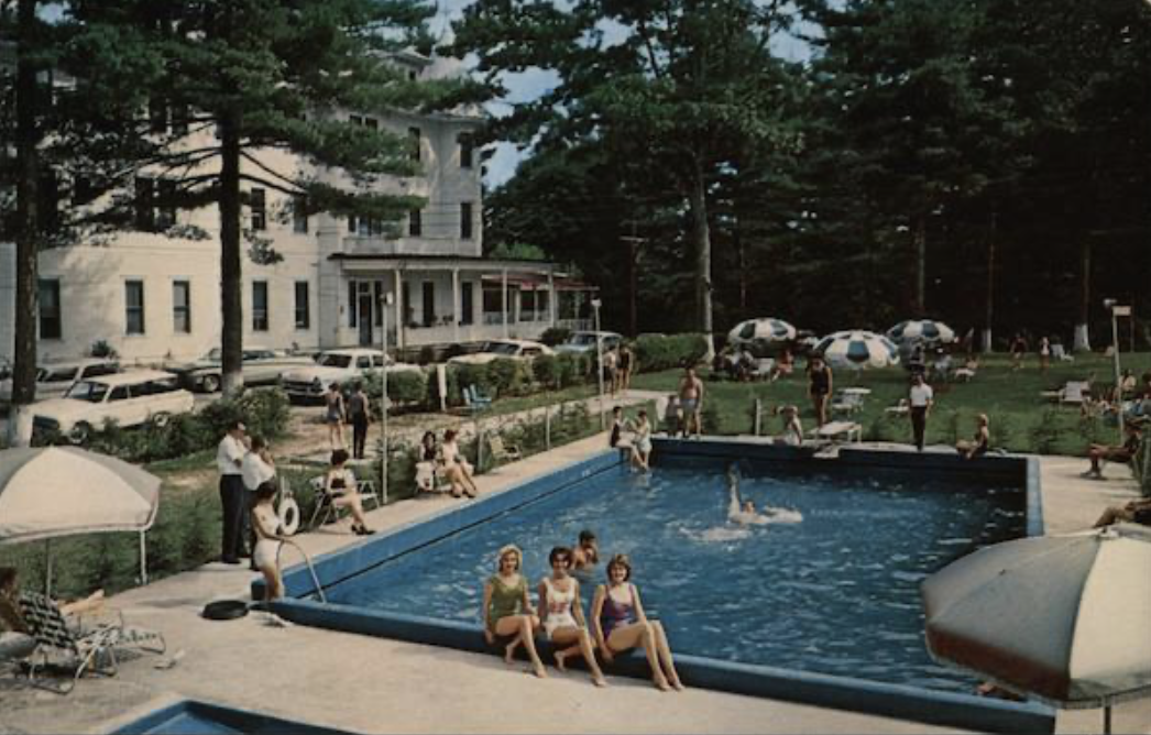People enjoying a sunny day at a public swimming pool surrounded by trees, with a large white house in the background. Some are sitting by the pool, others swimming or walking around. There are umbrellas and lounge chairs around the pool area.