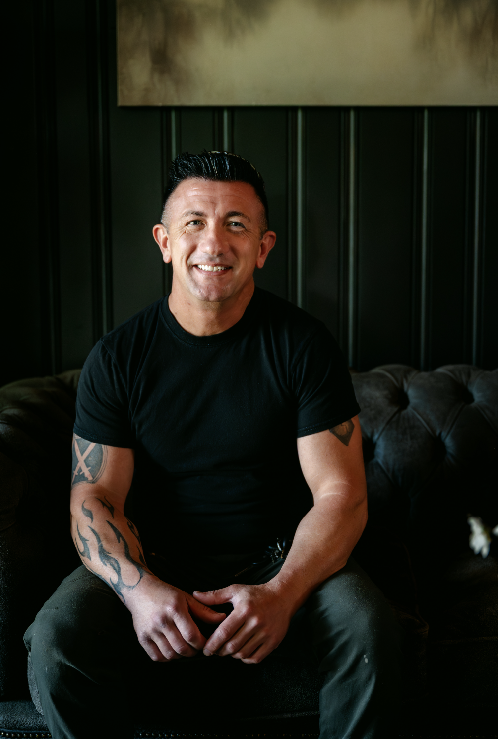 A smiling man with dark hair styled upward, wearing a black T-shirt, seated on a dark leather couch in a dimly lit room with dark green wall panels and a beige painting behind him.