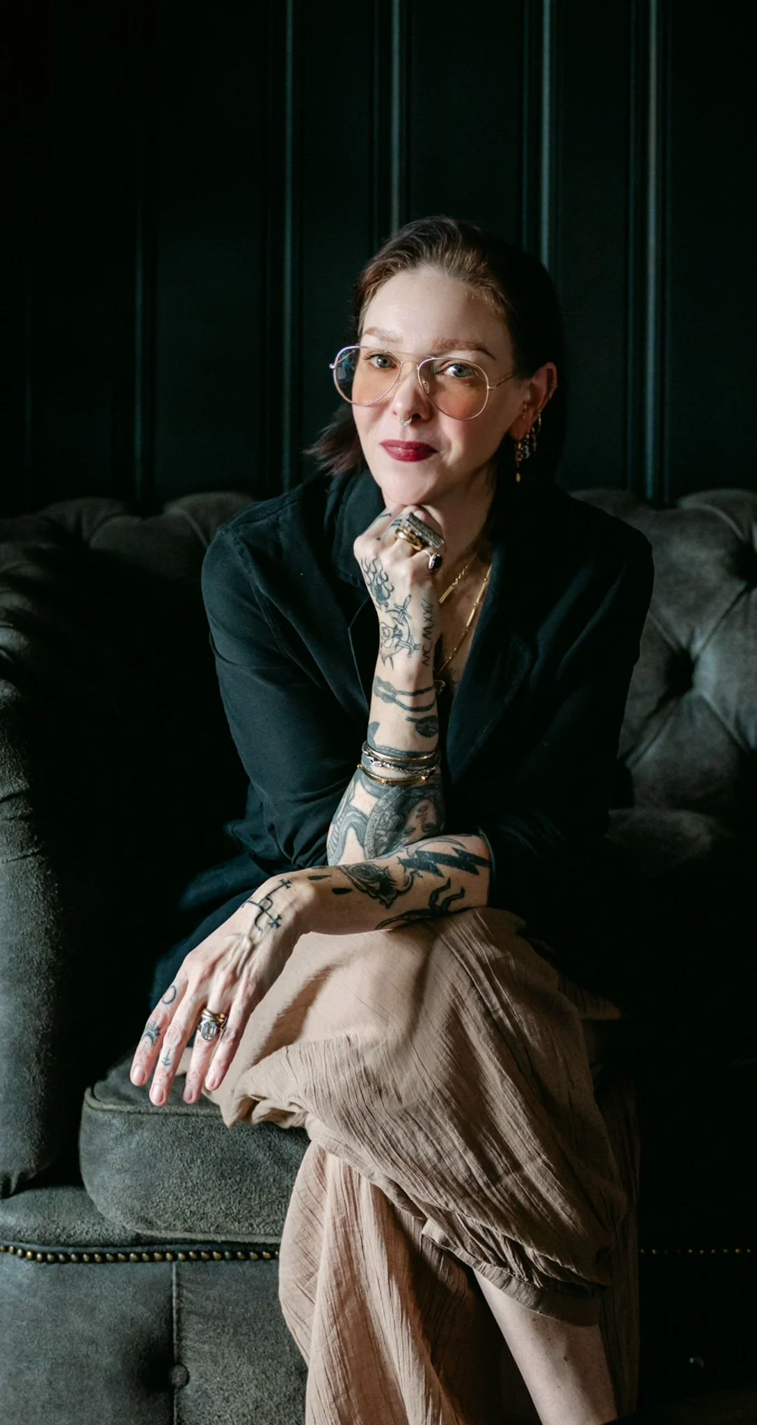 Woman with tattoos, glasses, and jewelry, sitting on a dark-colored sofa against a dark wooden paneled background.