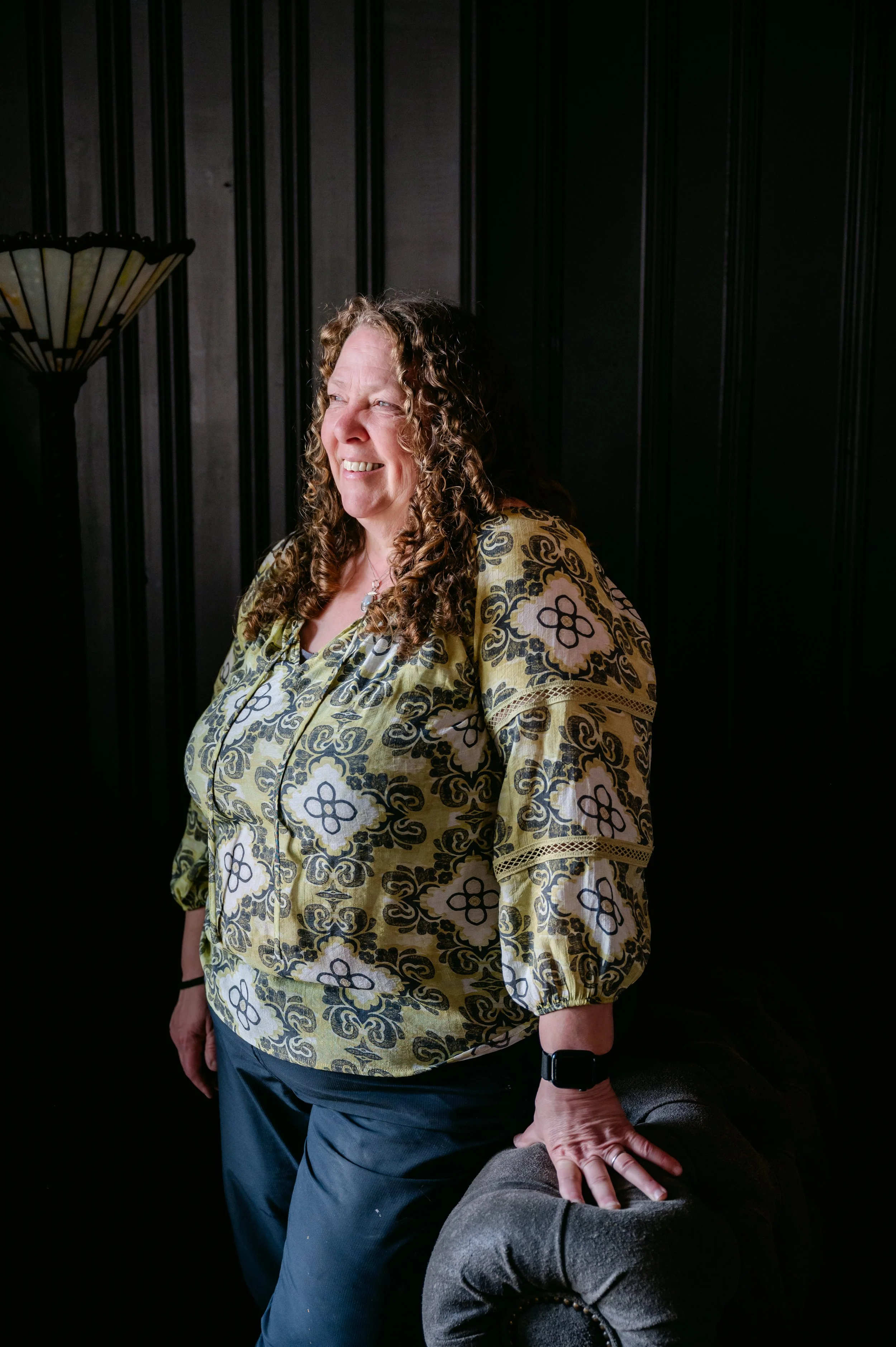 A woman with curly hair smiling, standing next to a dark-colored armchair in a room with black paneled walls and a decorative wall lamp.
