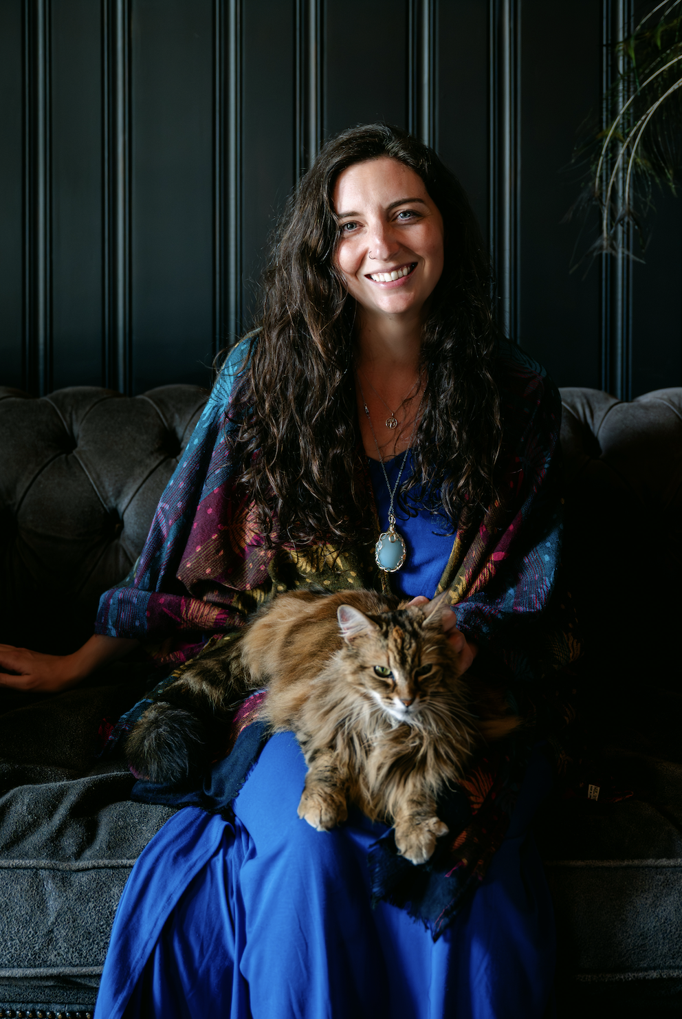 A woman with long dark wavy hair sitting on a dark sofa, smiling, holding a large cat with long fur.