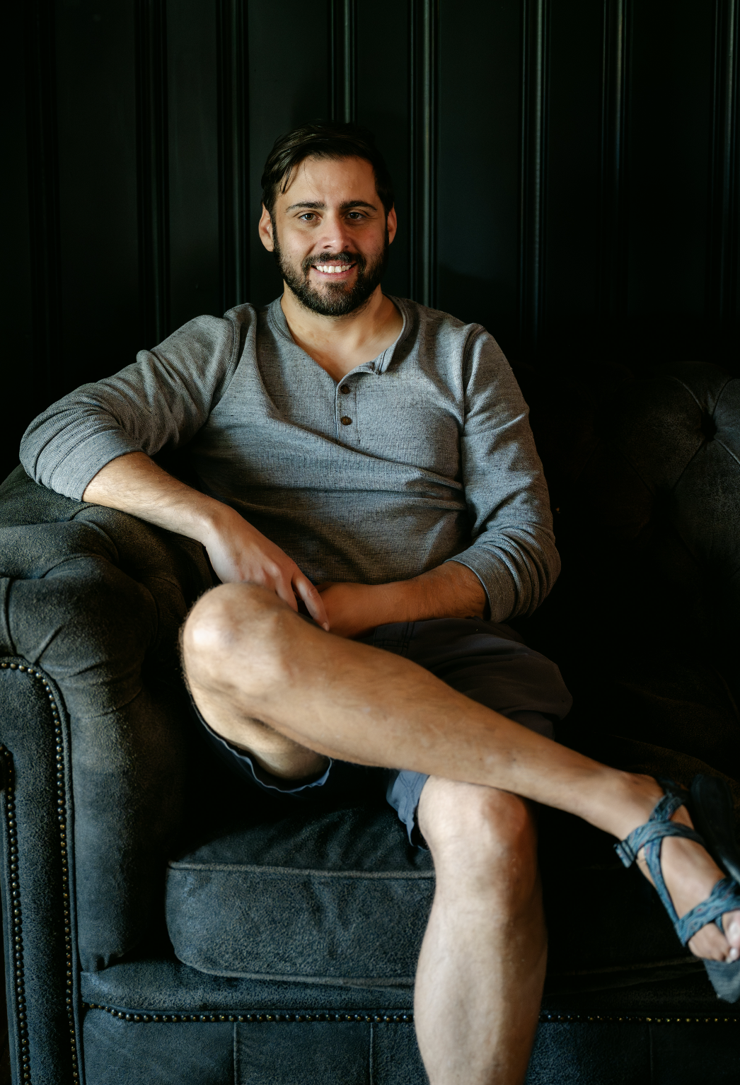 A man with dark hair and a beard, sitting on a black leather sofa with a dark wood paneled wall behind him, smiling at the camera, wearing a gray long-sleeve shirt and shorts.