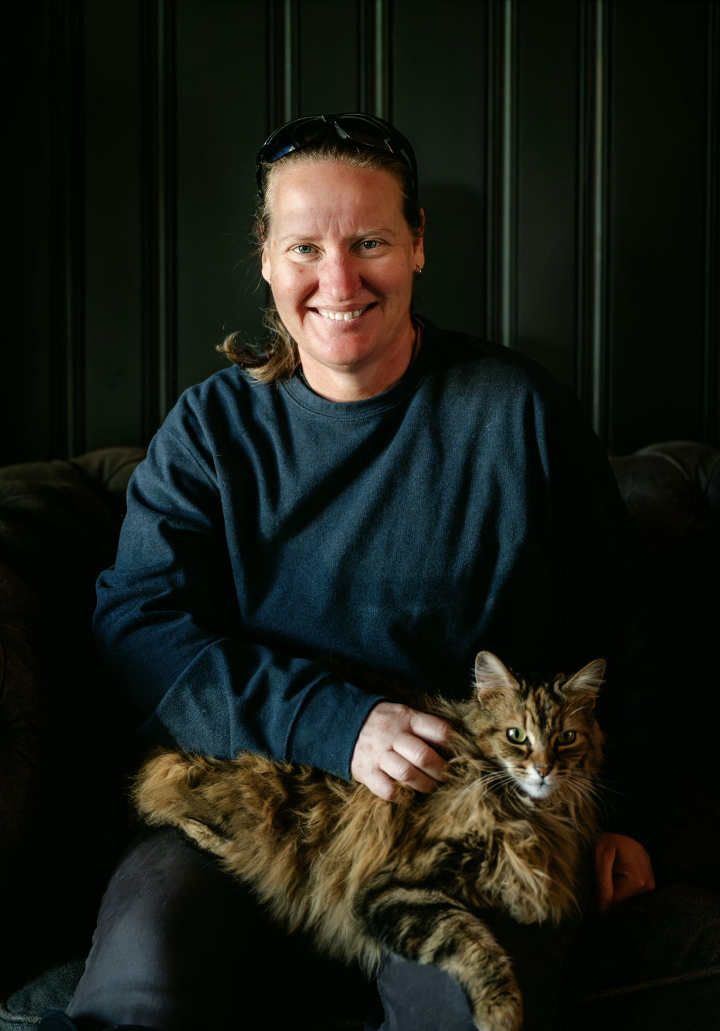 A woman with sunglasses on her head smiling and sitting on a dark-colored sofa in front of a dark green paneled wall, holding a long-haired tabby cat in her lap.