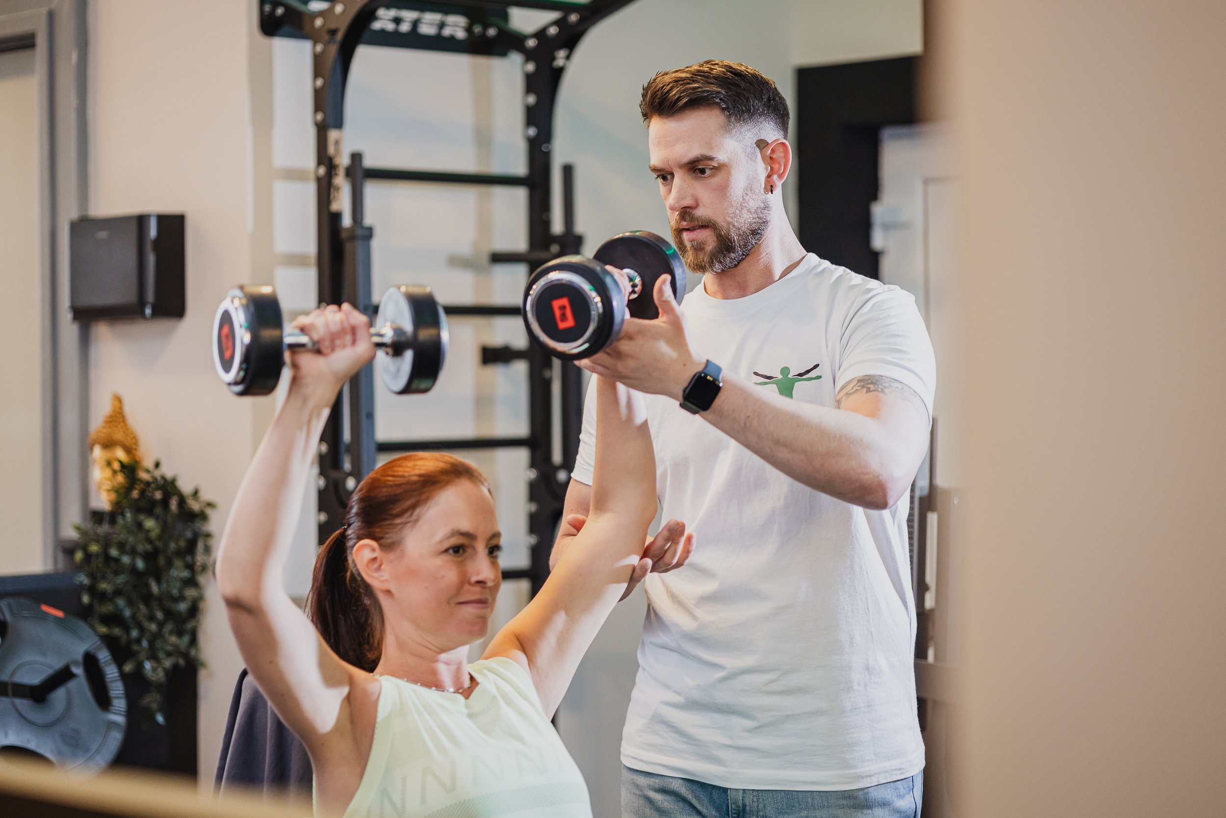 Ein Mann und eine Frau trainieren im Fitnessstudio. Der Mann hilft der Frau bei der Übung mit einer Hantel, während sie die Arme hebt.